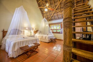 Habitación con dos camas, cada una con dosel, un banco de madera, ventilador de techo y una ventana con vitrales.