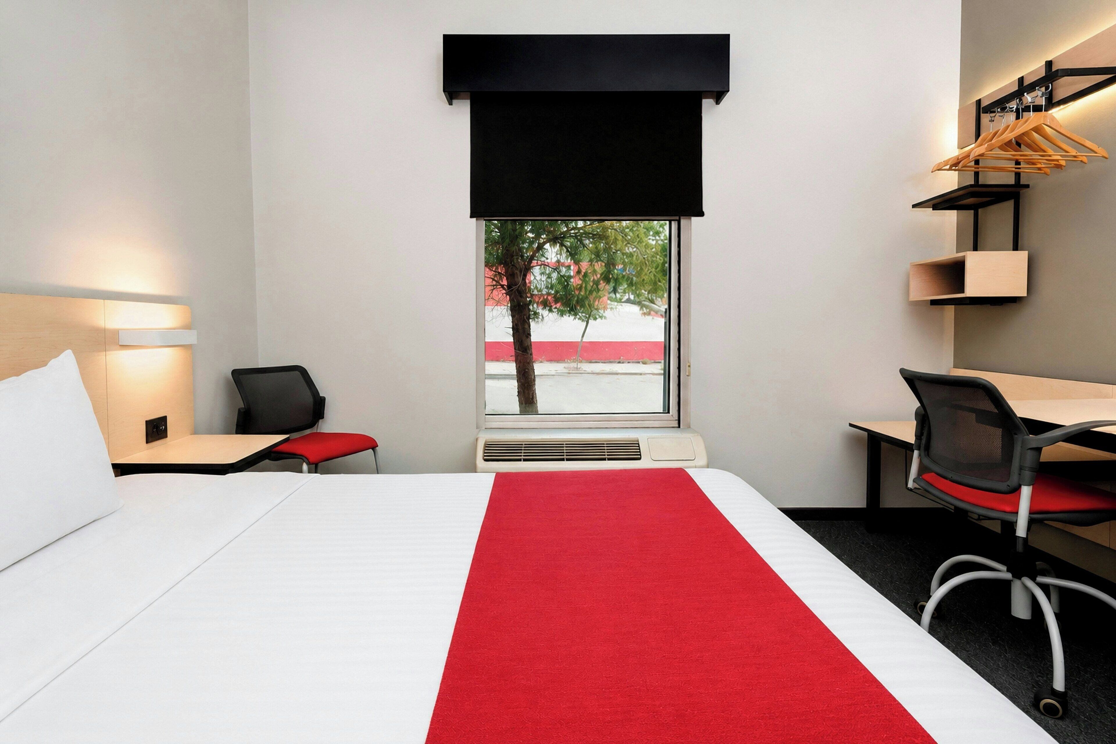 Ein Hotelzimmer mit Bett, Schreibtisch, Stuhl, Fenster mit Blick und einem an der Wand befestigten Regal mit Kleidung.