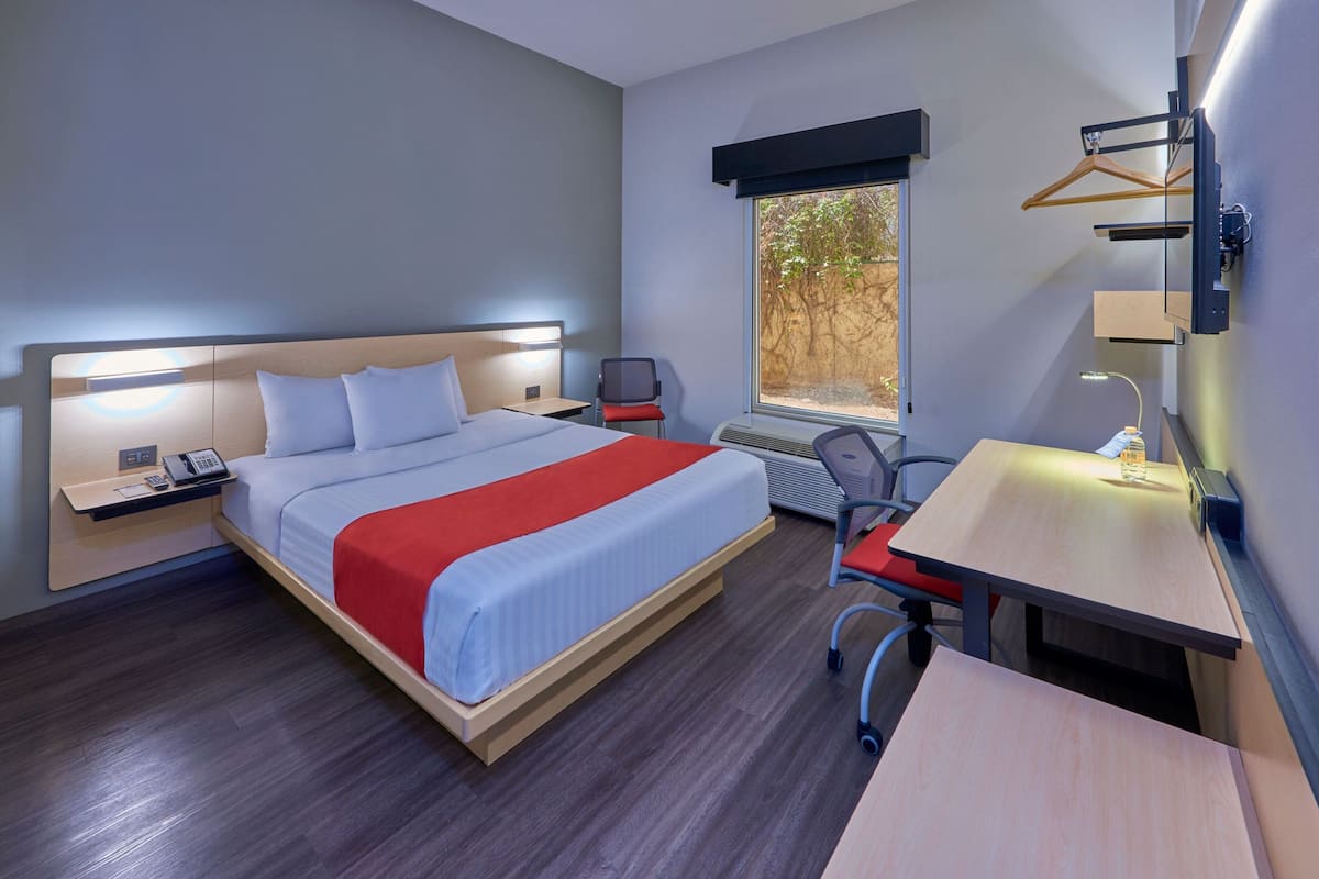 A hotel room with a bed, bedside table, desk, chair, and a window with a view of trees.