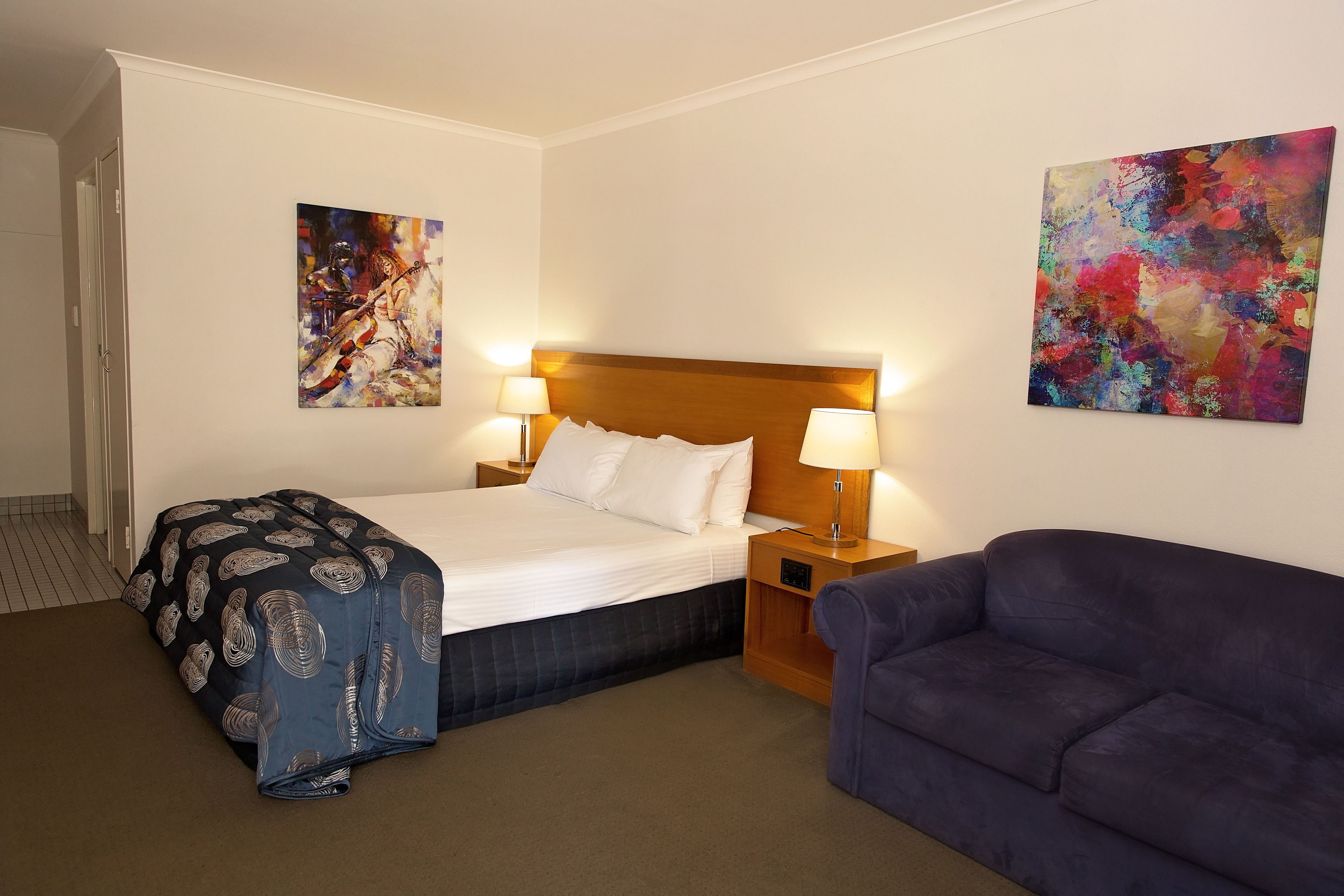 Habitación de hotel con cama, dos lámparas de noche, un sofá azul y dos cuadros abstractos en las paredes.