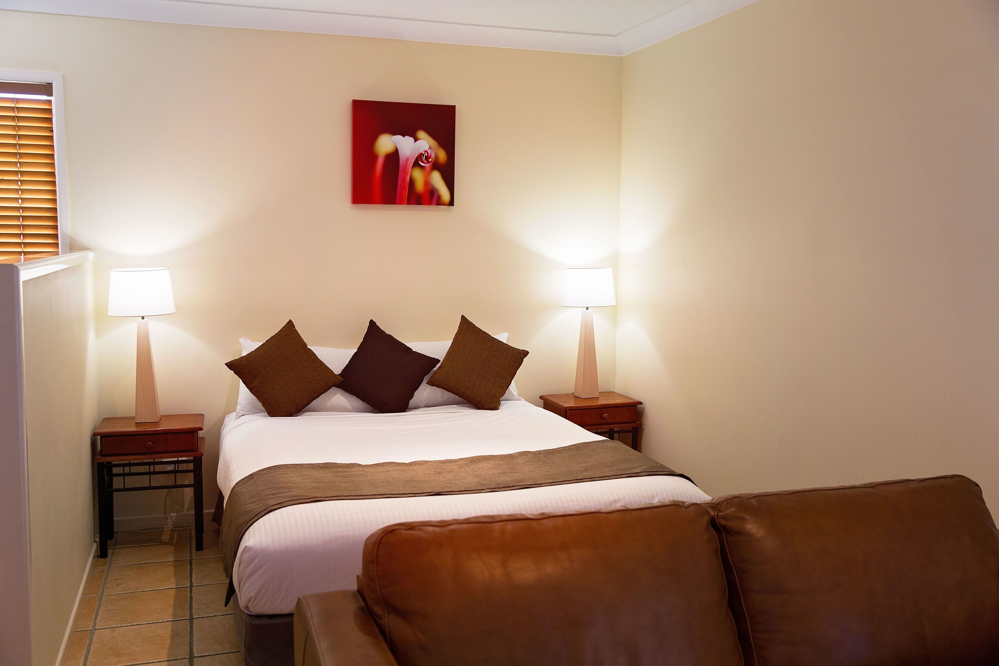 A neatly arranged hotel room with a bed, two bedside tables with lamps, a brown leather chair, and a red abstract painting on the wall.