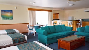 A hotel room with two beds, a teal sofa, a wooden coffee table, a dining area with chairs, and a kitchen area. - White Lace Motor Inn (West Mackay)