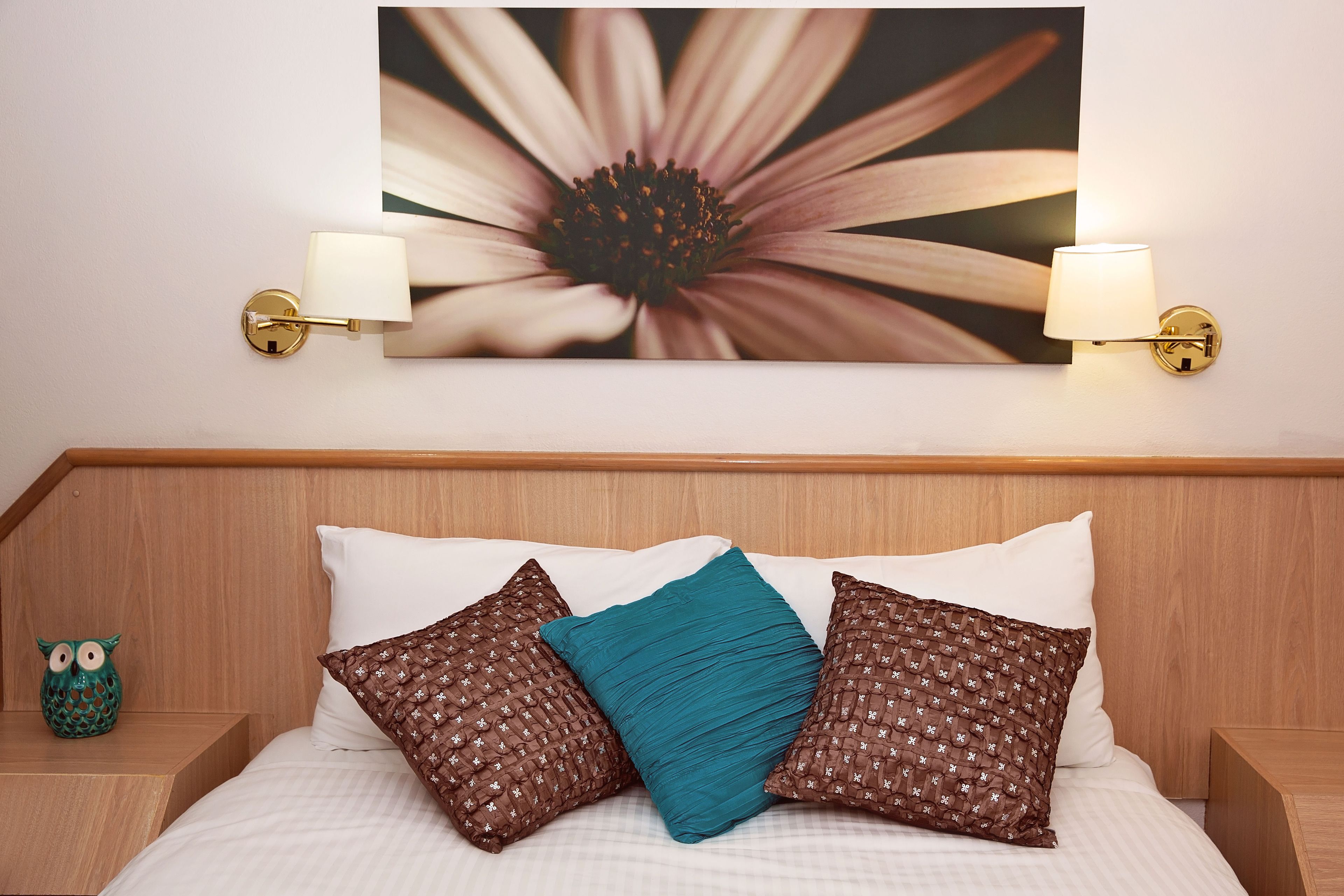 Un lit bien fait, agrémenté d’oreillers à motifs, une lampe fixée au mur et, au-dessus, un grand tableau représentant des fleurs.