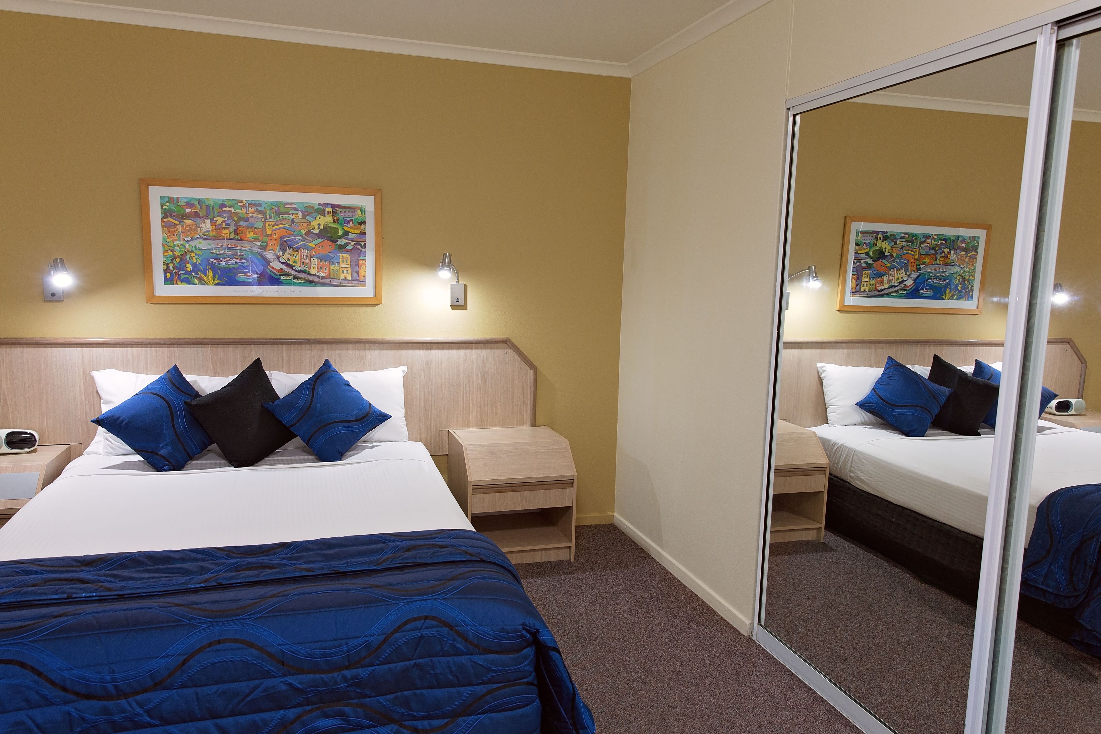 A hotel room with a bed, a nightstand, a bedside table, a mirror, and a painting on the wall.