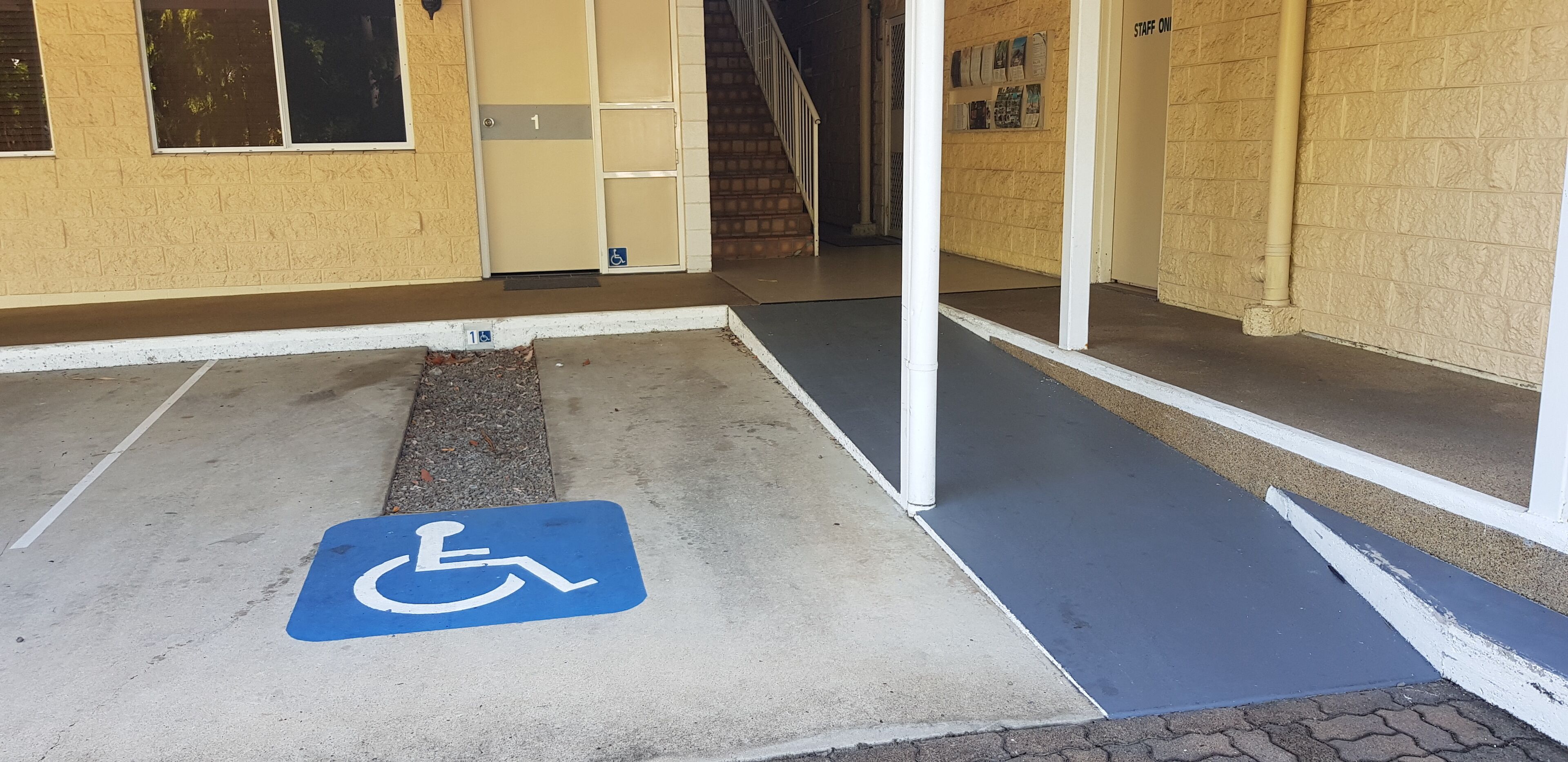 Plaza de aparcamiento accesible para sillas de ruedas señalizada con un letrero azul, una entrada a edificio con rampa y una escalera que conduce al interior.