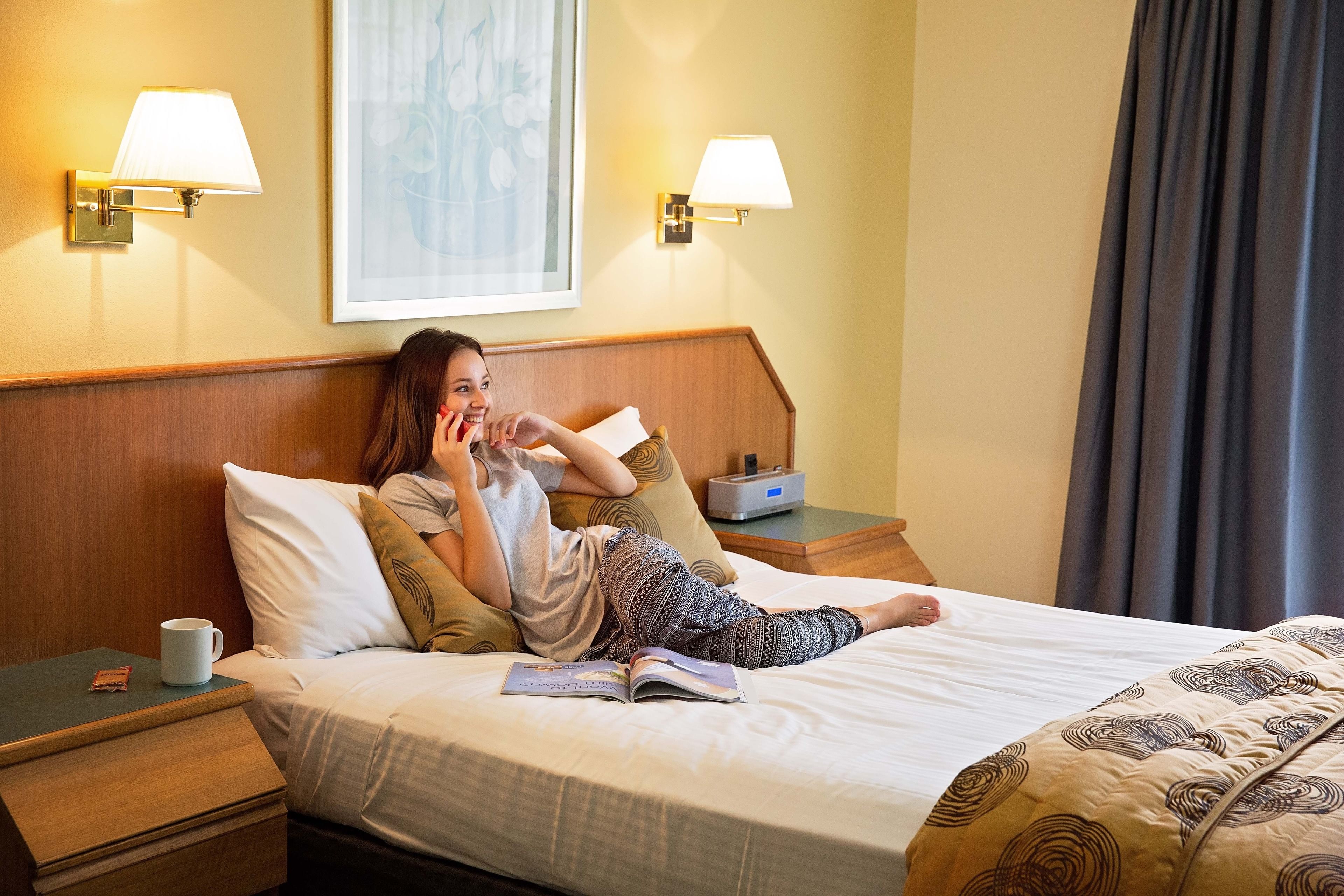 A hotel room with a bed, a bedside table with a lamp, a bedside table with a cup, and a wall-mounted lamp.