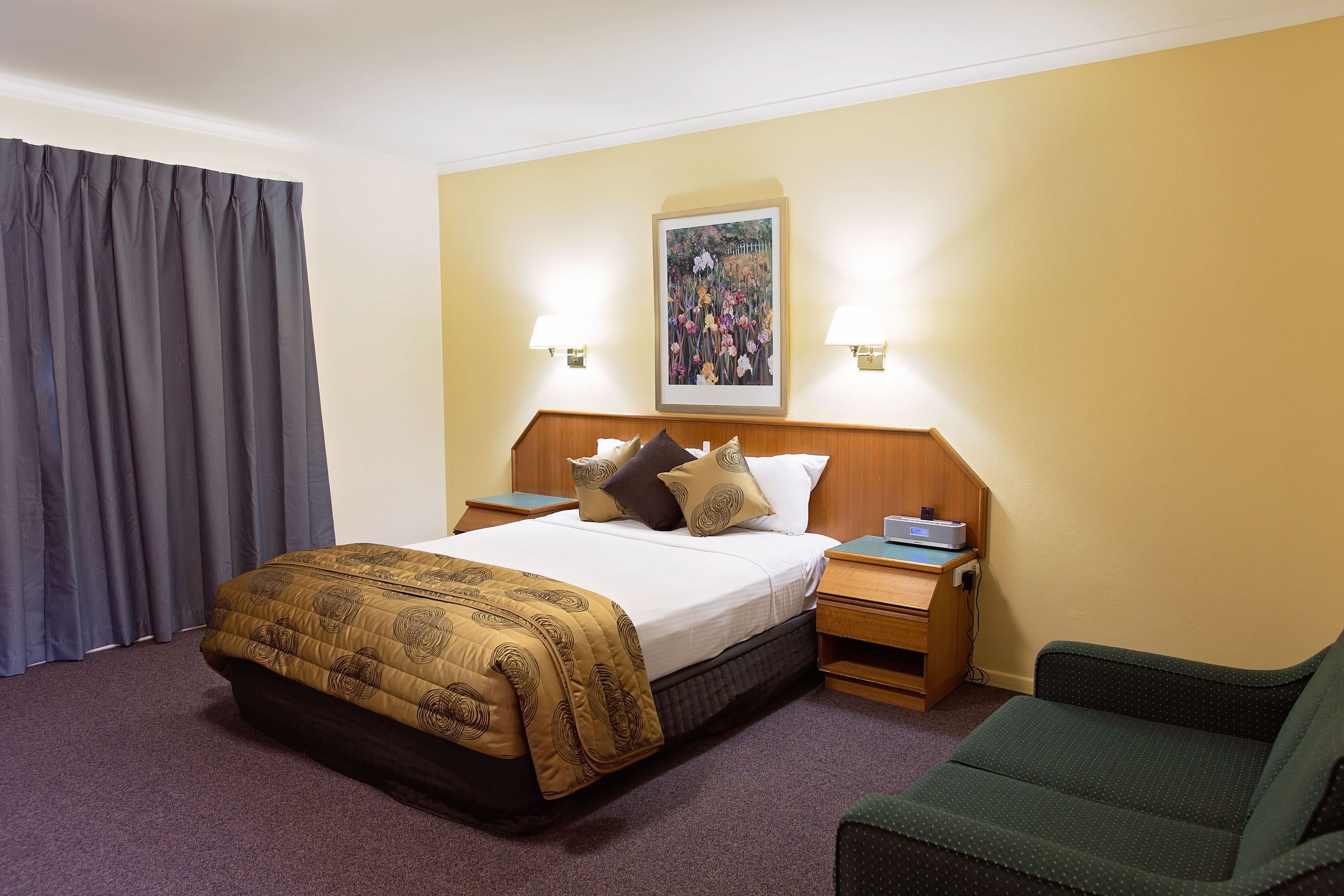 Ein Hotelzimmer mit einem Bett, zwei Nachttischen, einem Sessel, einem Bild an der Wand und einem Fenster mit Vorhängen.