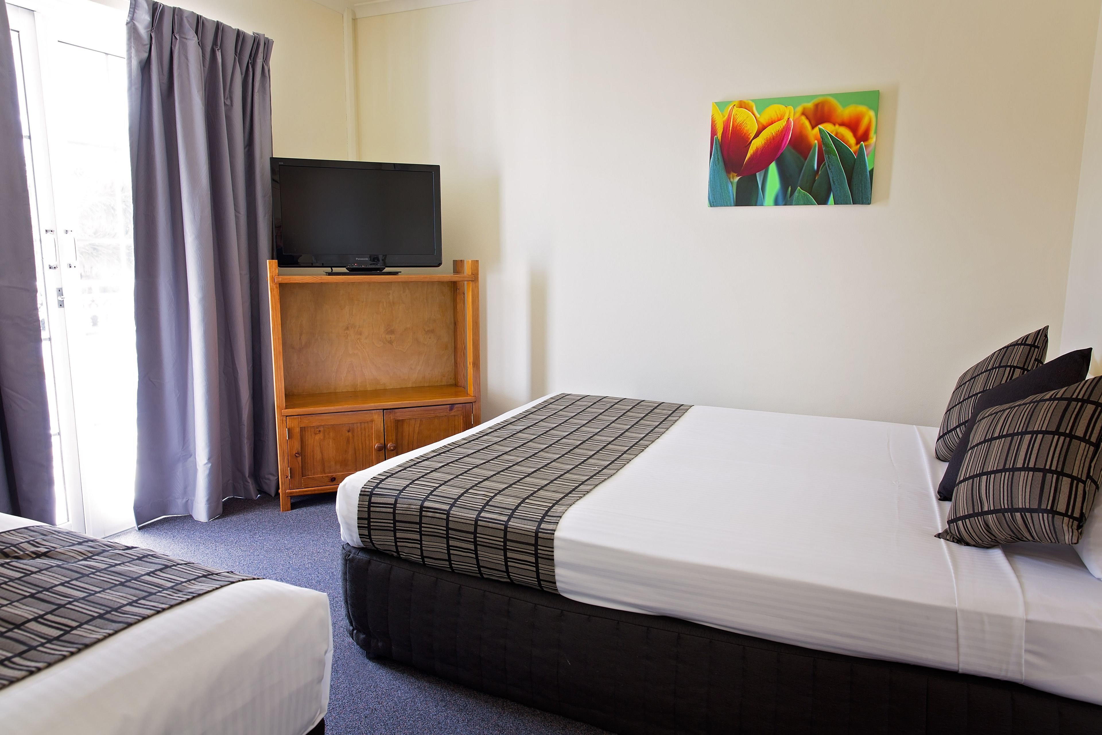 A hotel room with a bed, a TV on a wooden stand, a painting of flowers on the wall, and a window with curtains.