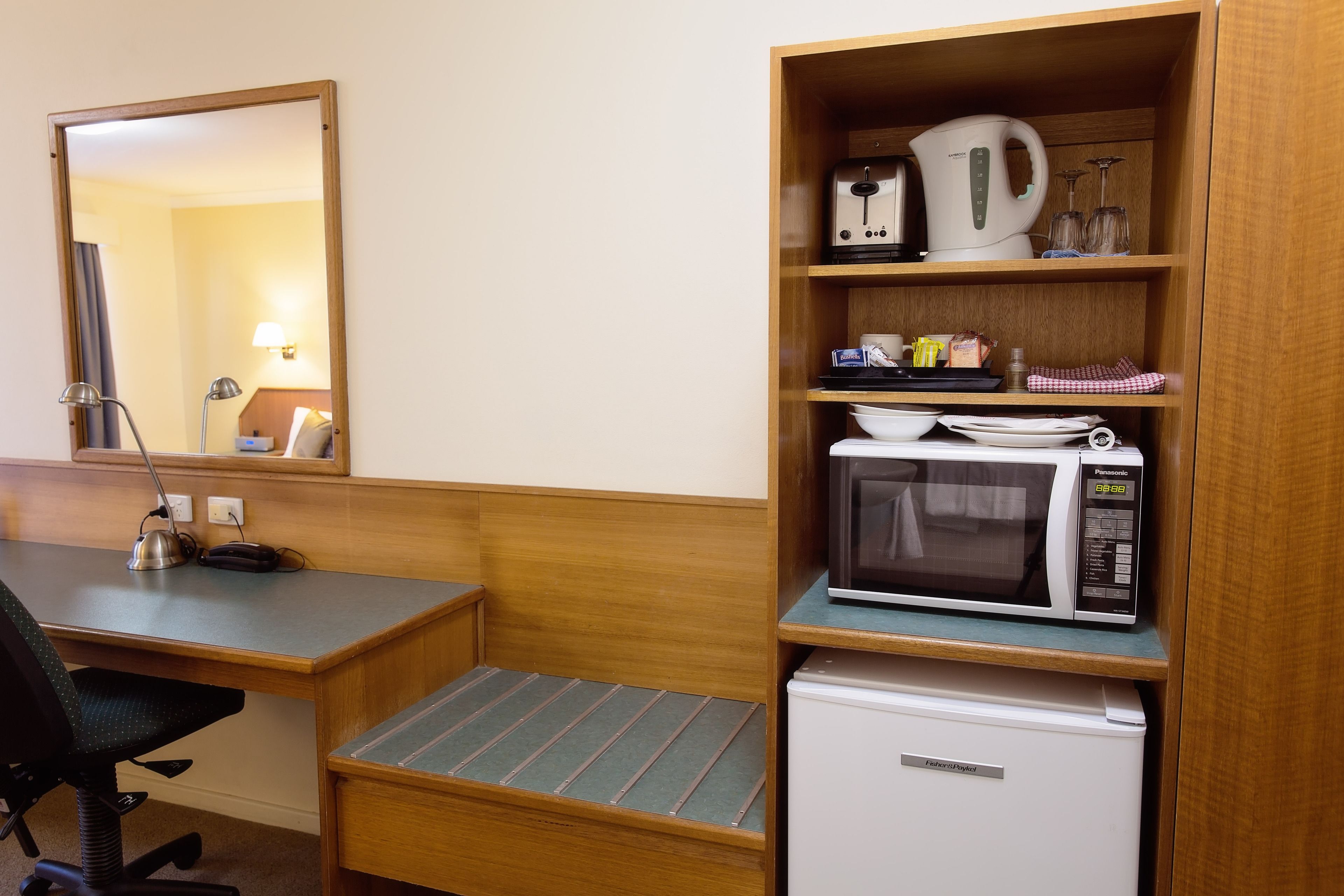 Une chambre d’hôtel compacte comprenant un bureau en bois, un petit réfrigérateur, un four à micro-ondes, une bouilloire et un miroir.