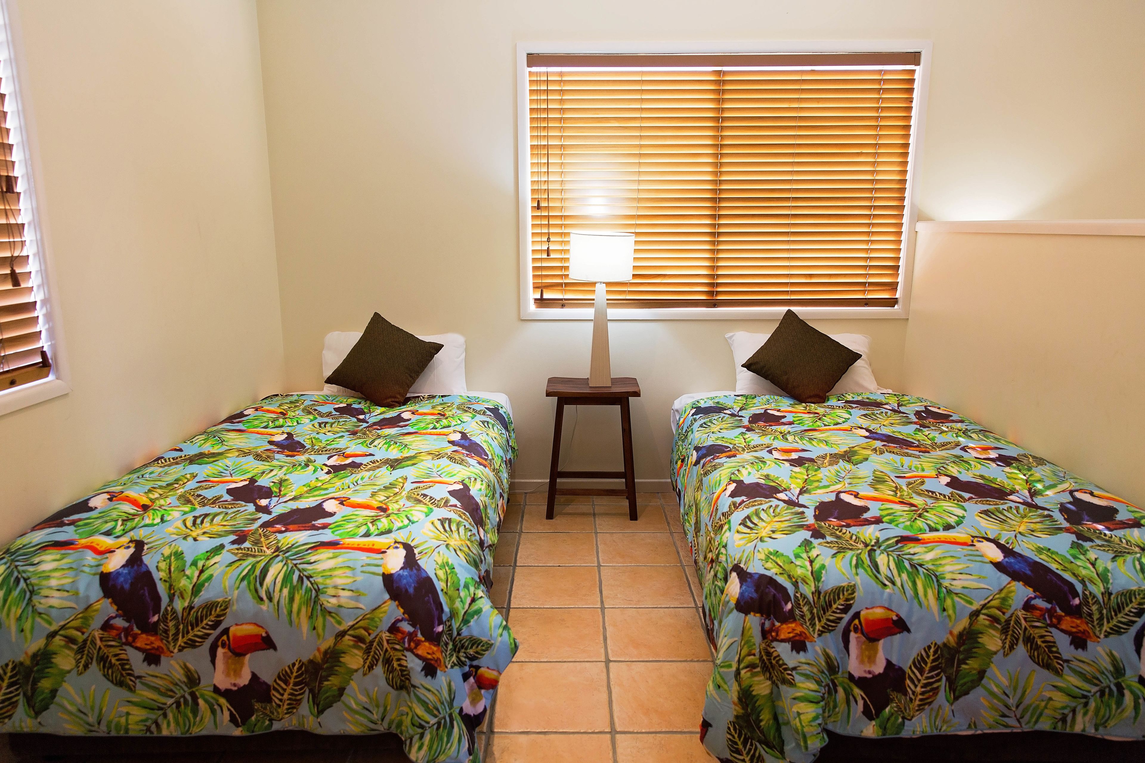Habitación con dos camas, cada una con colcha de colores que muestra tucanes y hojas, una mesita y una ventana con persianas.