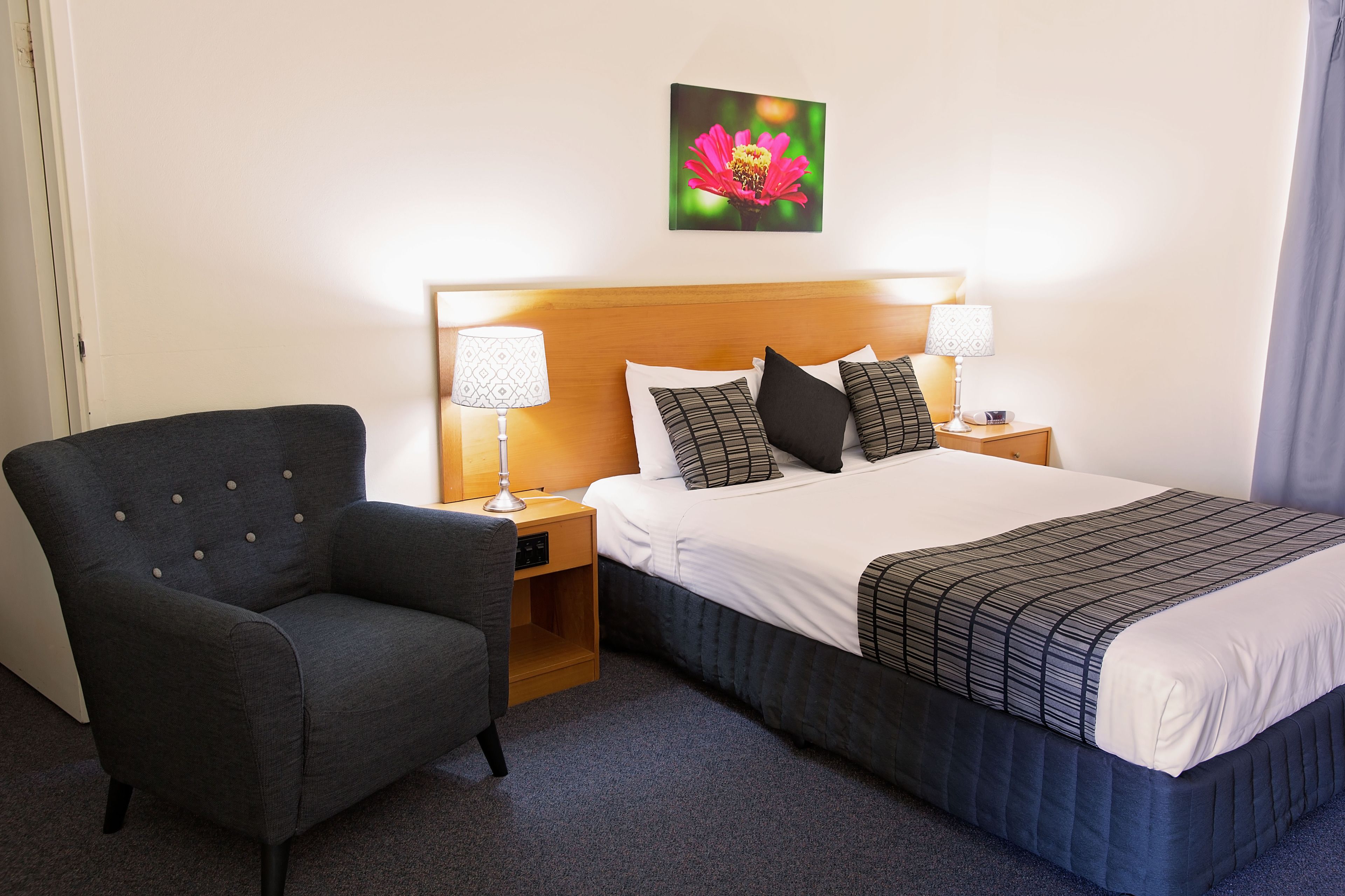 A hotel room with a bed, two bedside tables with lamps, a chair, and a wall-mounted picture of a flower.