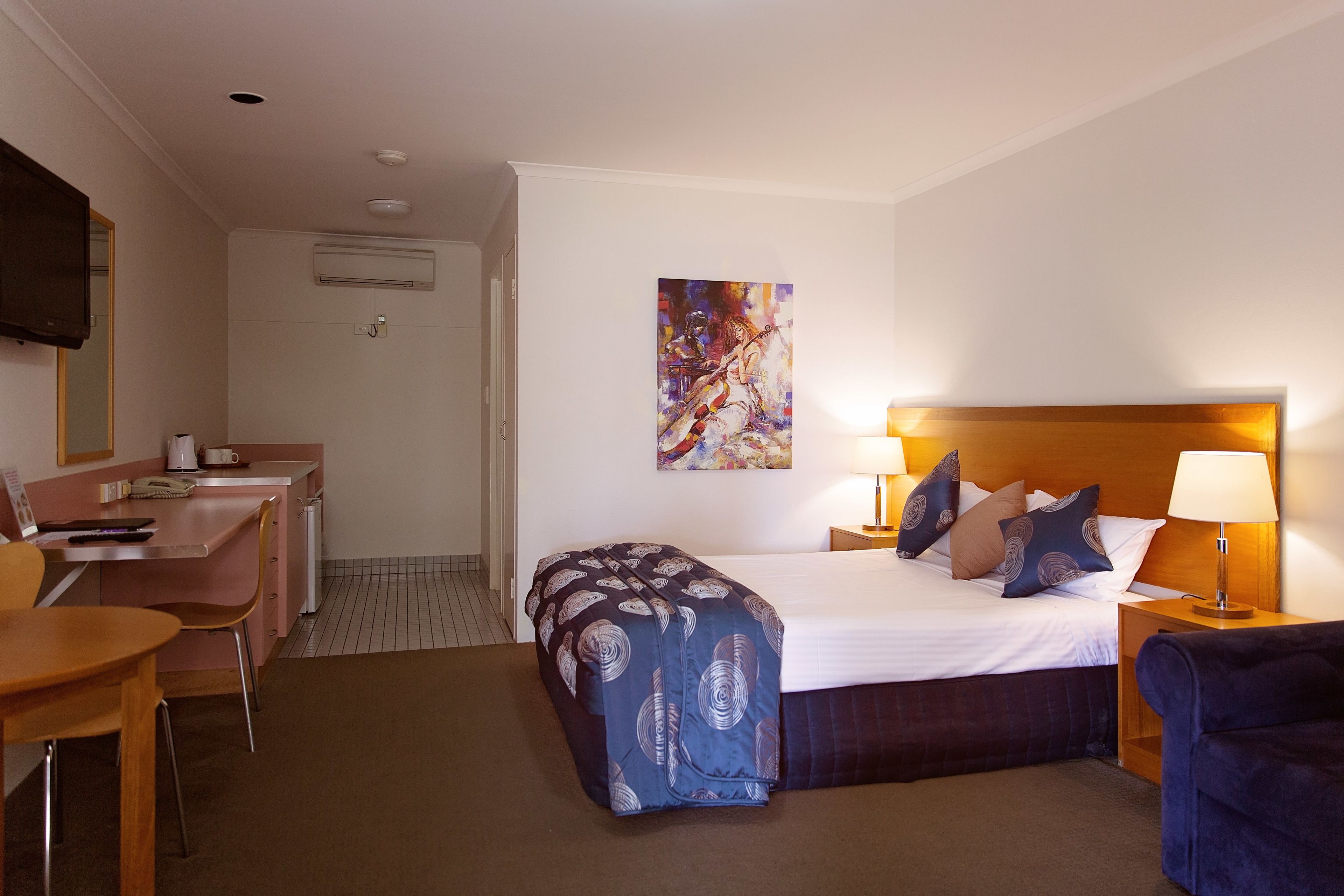A hotel room with a bed, bedside table, lamp, chair, desk, and a painting on the wall.