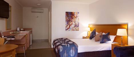 A hotel room with a bed, bedside table, lamp, chair, desk, and a painting on the wall.