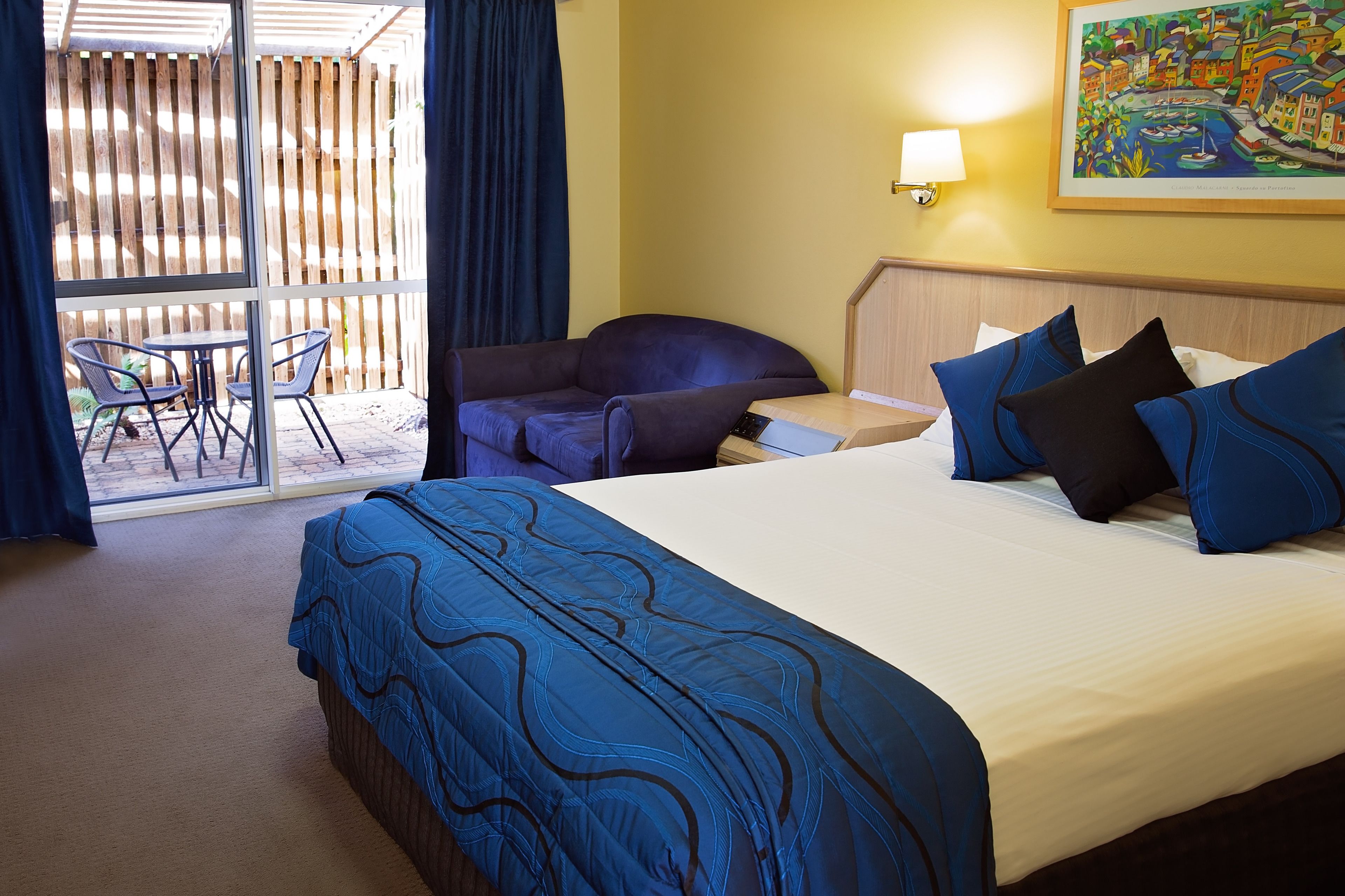 A hotel room with a large bed, a sofa, a small table, and a chair. There is a balcony with a view of the outside.