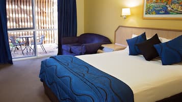 A hotel room with a large bed, a sofa, a small table, and a chair. There is a balcony with a view of the outside.