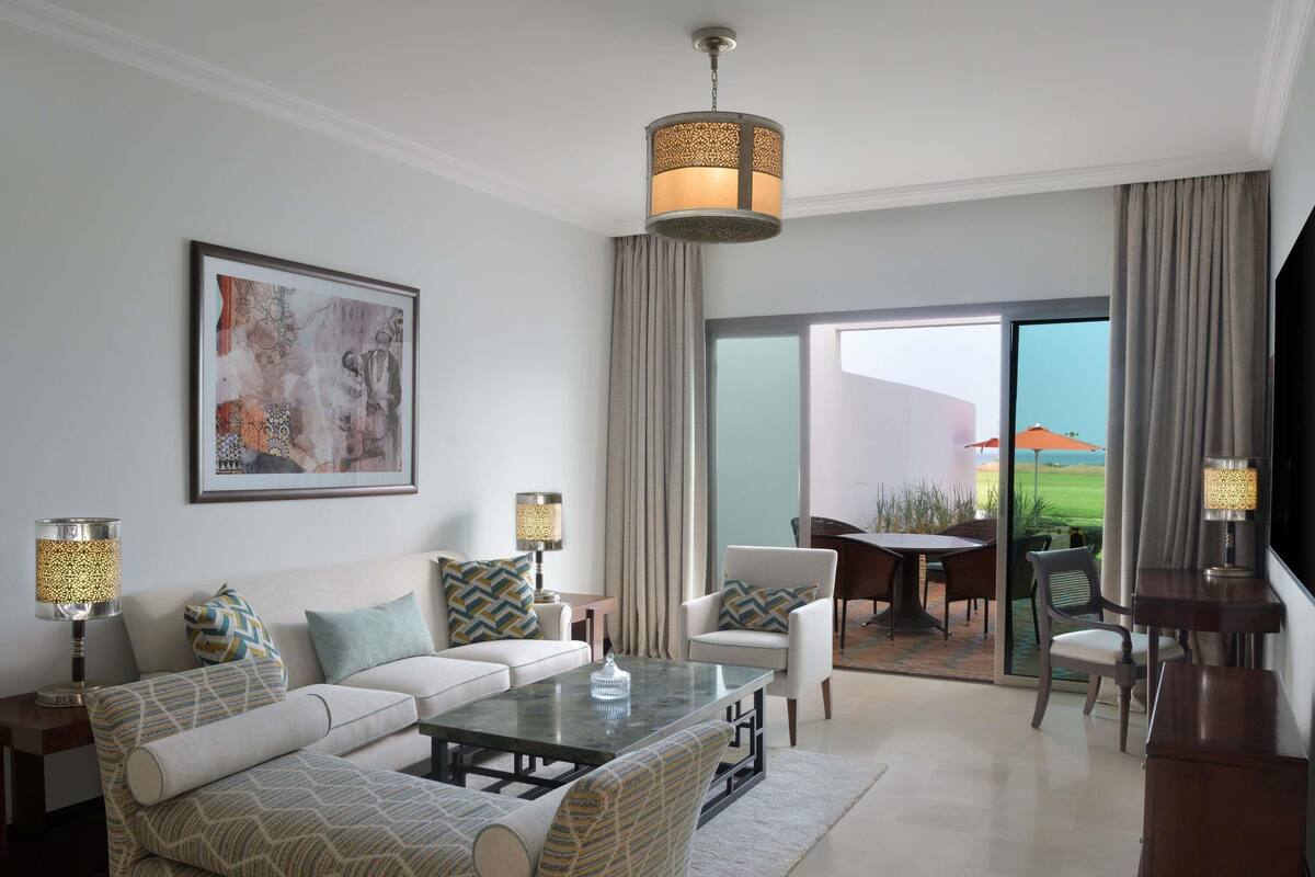 A modern living room with a sofa, coffee table, and lamps, a dining area with chairs, and a large window with a view.