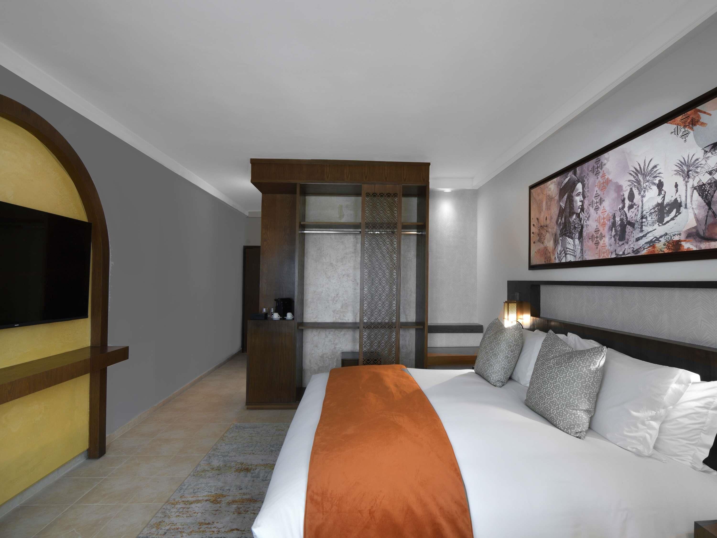 A hotel room with a large bed, a television mounted on the wall, a painting above the bed, and a wooden wardrobe.