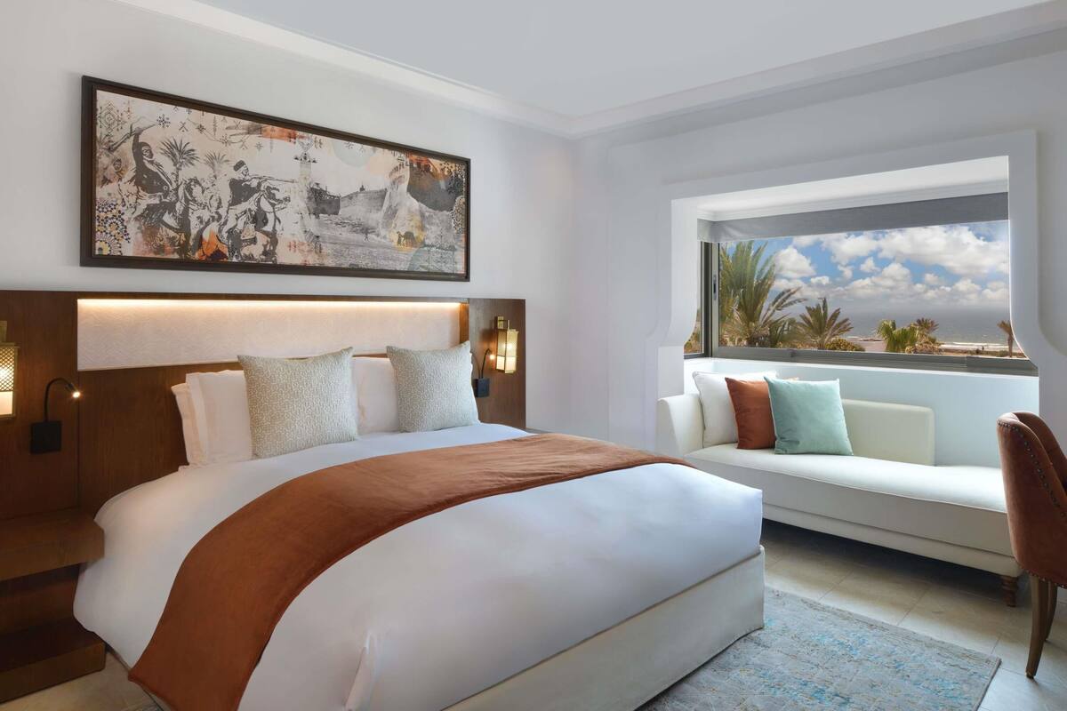 A modern hotel room with a large bed, a sofa, a view of the beach, and a large framed artwork on the wall.
