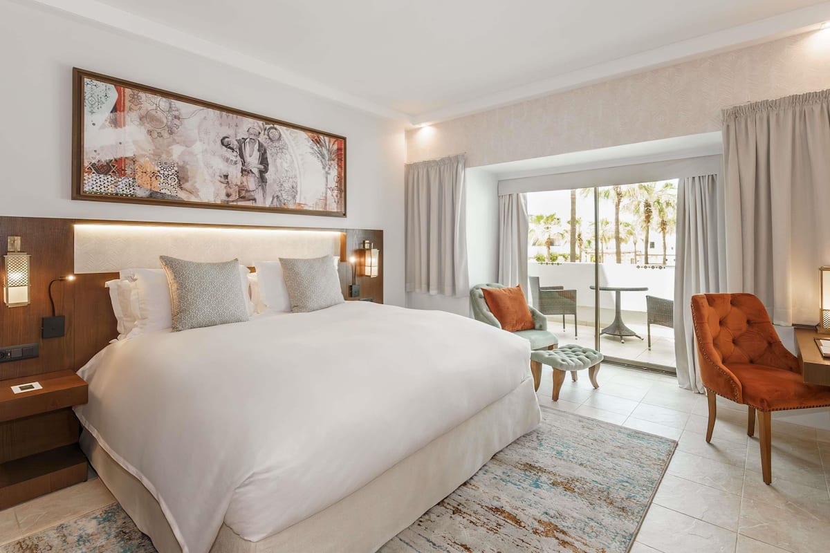 A hotel room with a large bed, a painting above the bed, a balcony with chairs, and a view of palm trees.