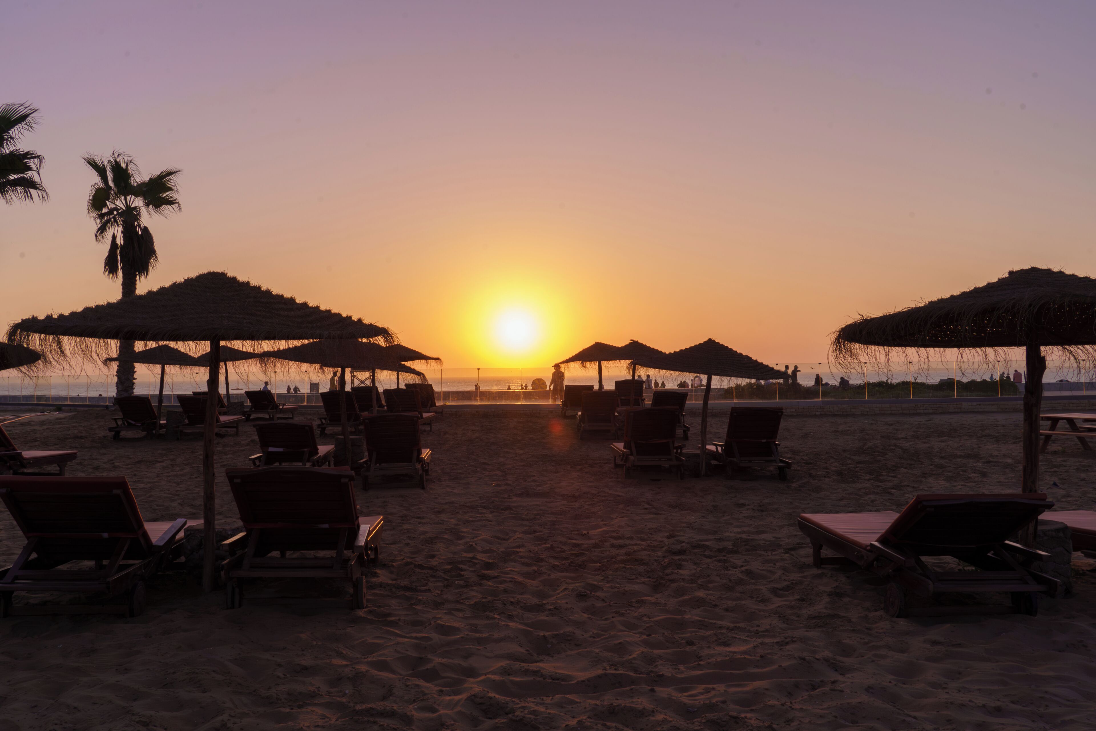 A beach with sun loungers and thatched umbrellas under a sunset sky.