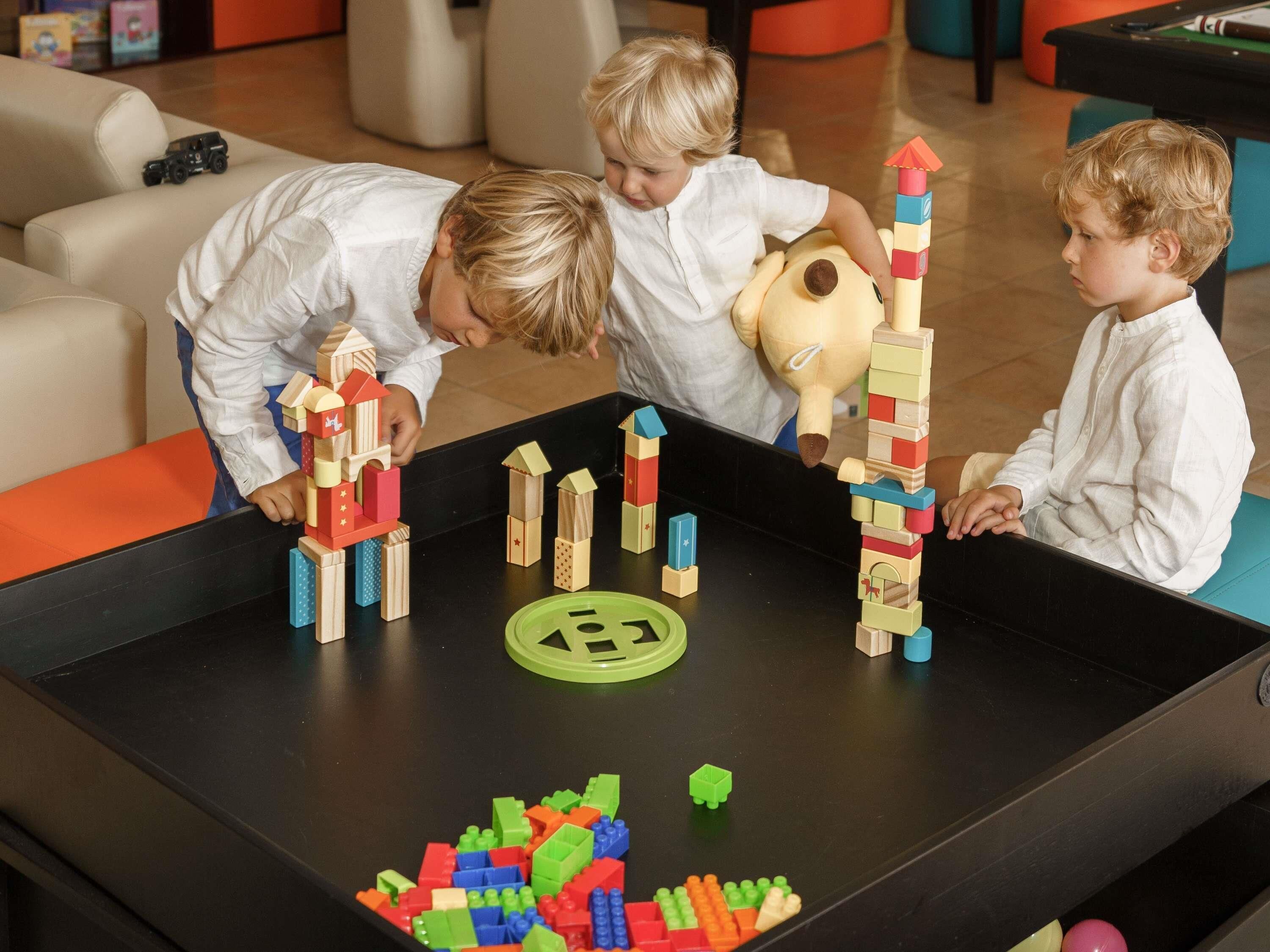 A group of children playing with building blocks on a table in a room with a pool table and comfortable seating in the background.