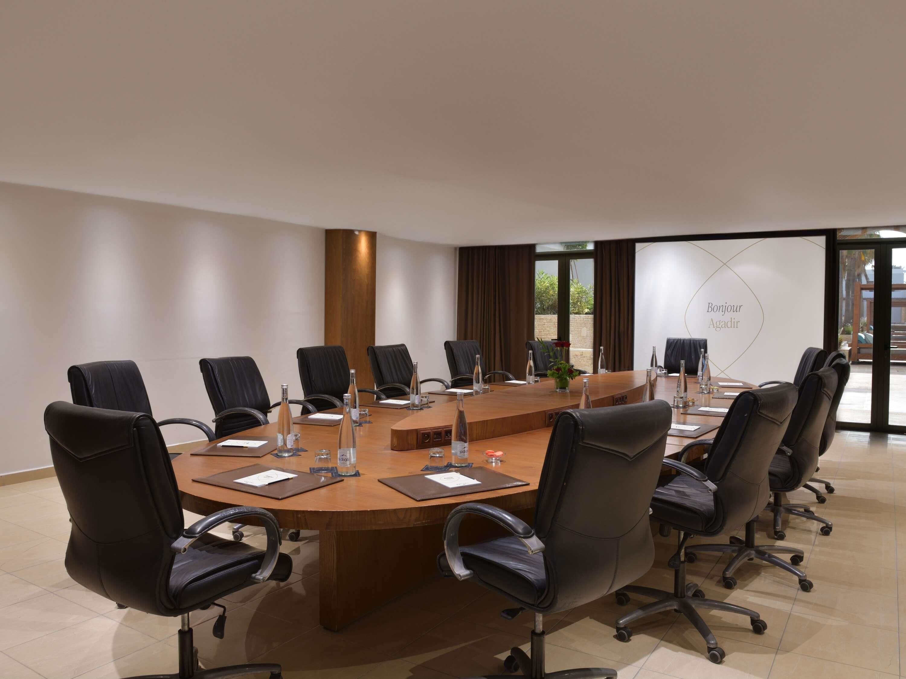 A conference room with a large oval table, black leather chairs, water bottles, and a 'Bonjour Agadir' sign.