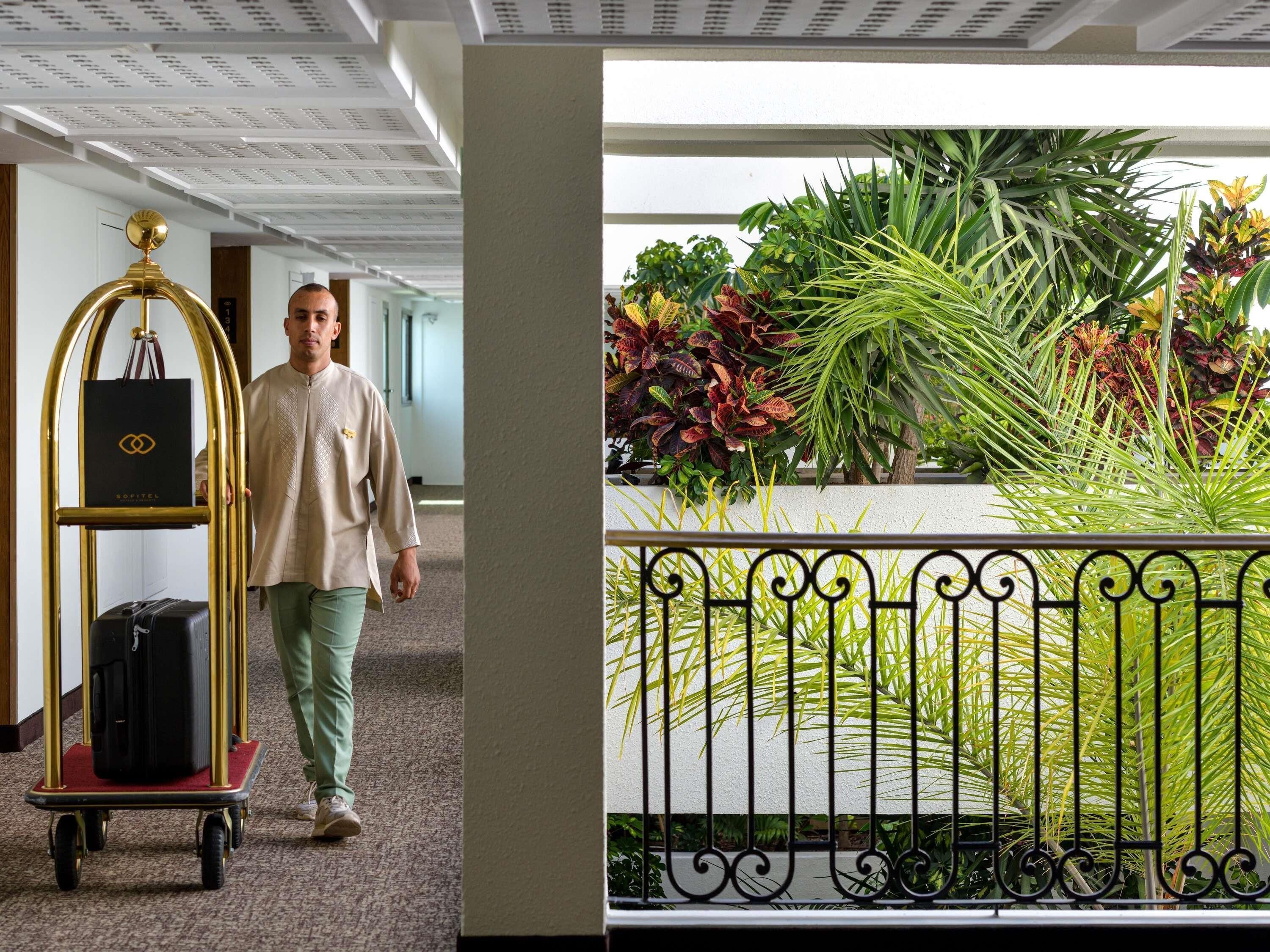 A man walking past a luggage cart with a black suitcase and a gold bag, leading to a room with a white ceiling and a view of a garden with various plants.