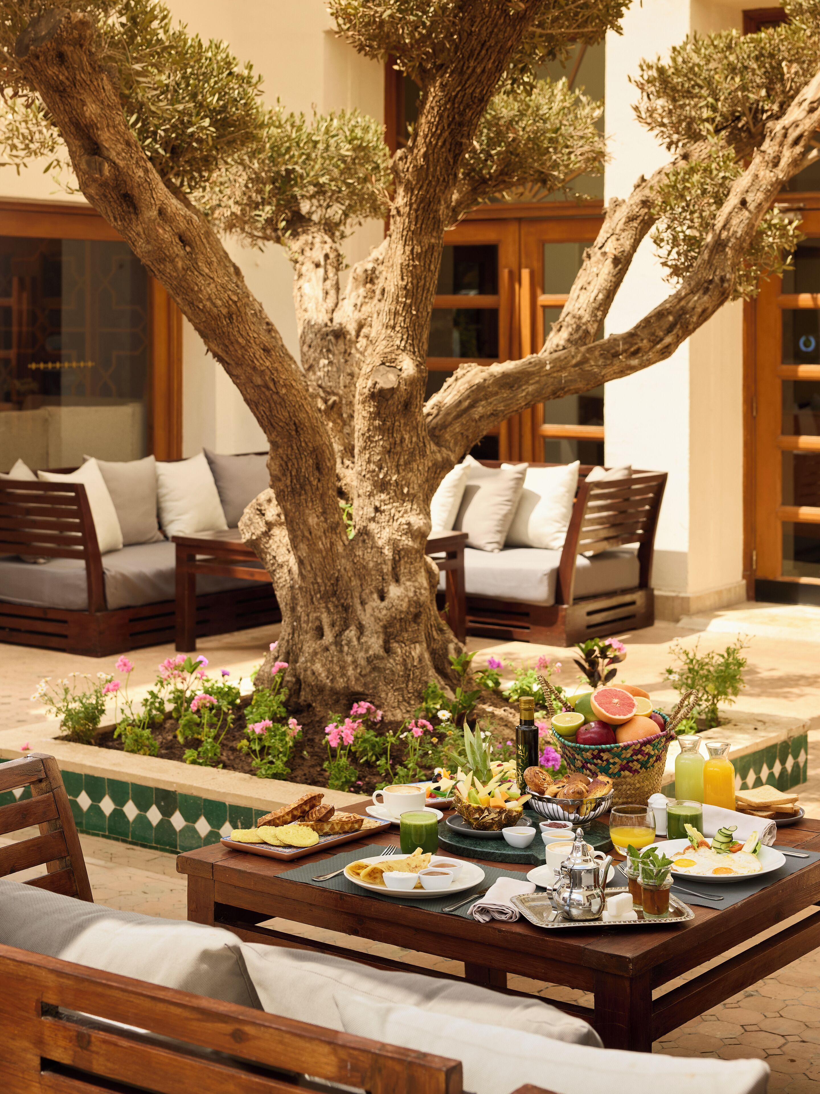 Un comedor con un gran árbol, asientos al aire libre y diversas opciones de desayuno sobre la mesa.