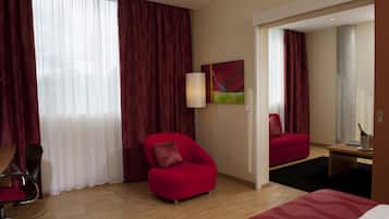 A hotel room with a red armchair, a bed, a desk with a lamp, and a large mirror.