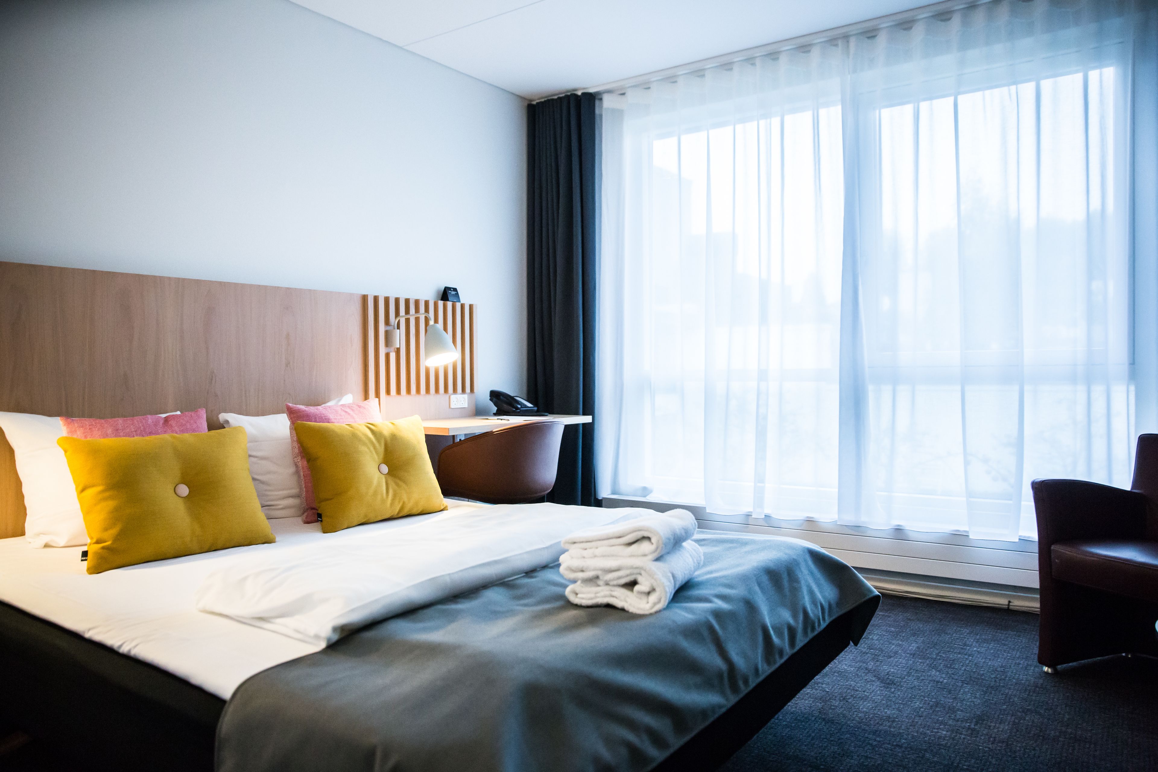 Quarto de hotel com cama, escrivaninha, cadeira e uma grande janela com cortinas finas.