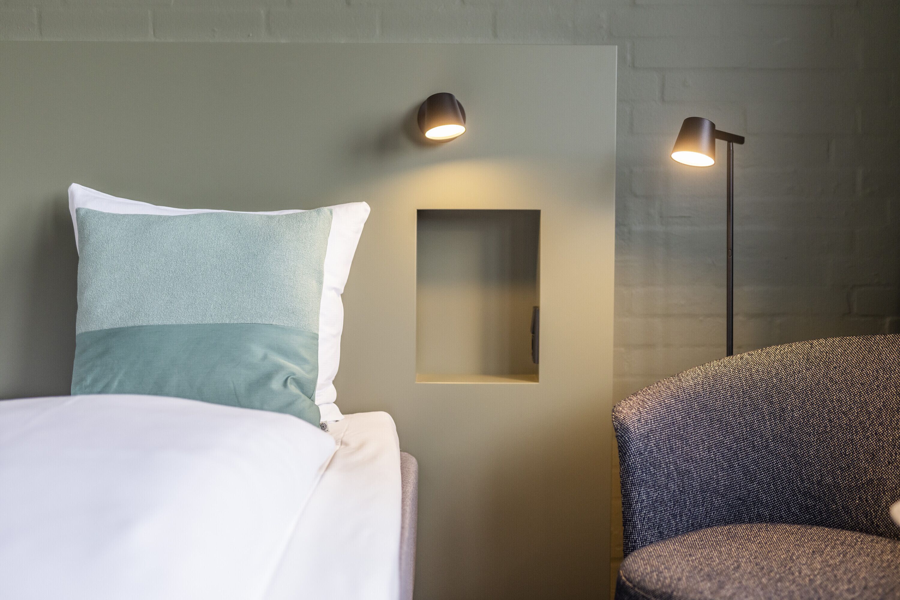 Una cama bien hecha con un almohadón, un espejo rectangular pequeño en la pared y una silla con lámpara.