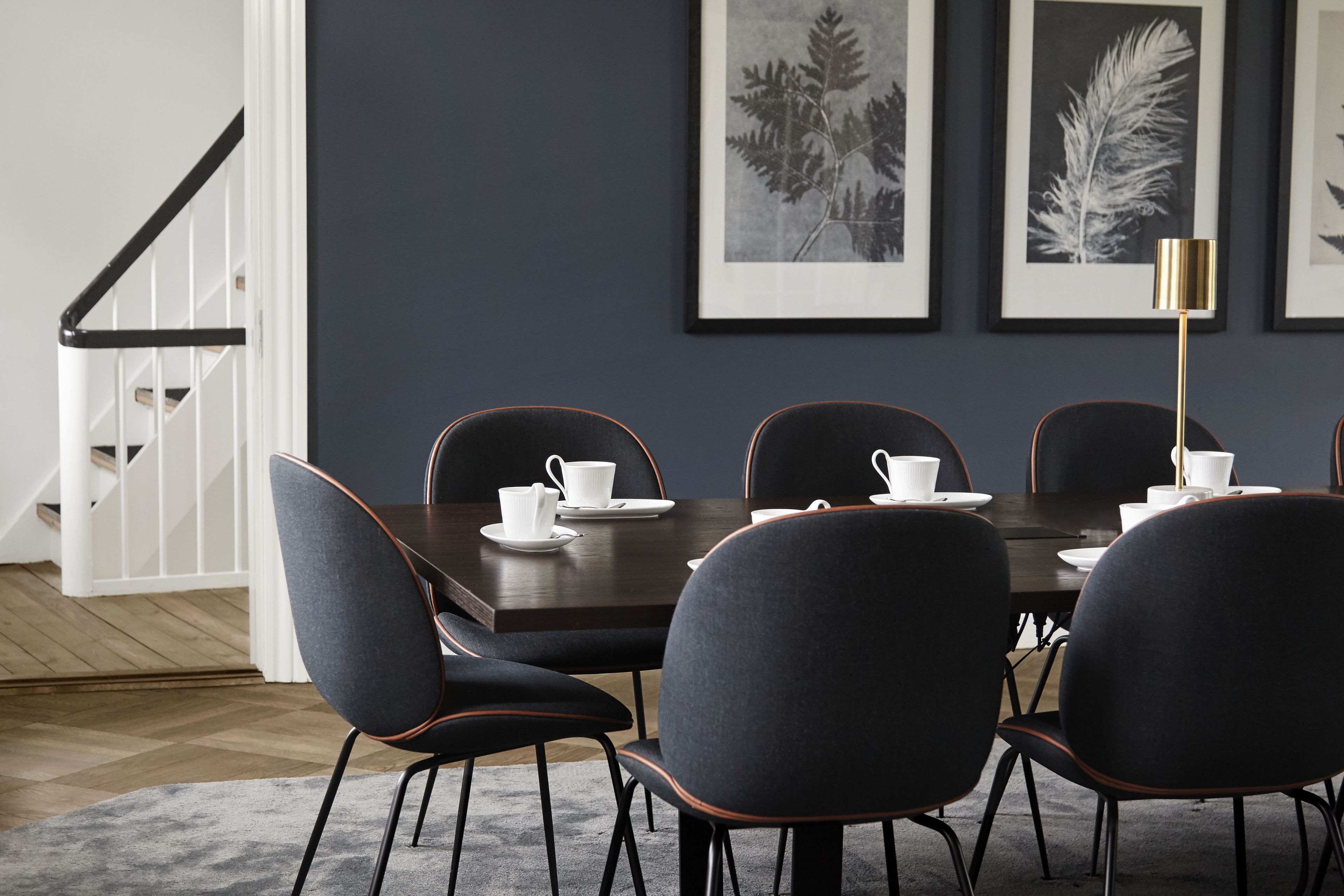 A dining area with a dark wooden table, six chairs with black upholstery and orange trim, and framed botanical prints on the wall.