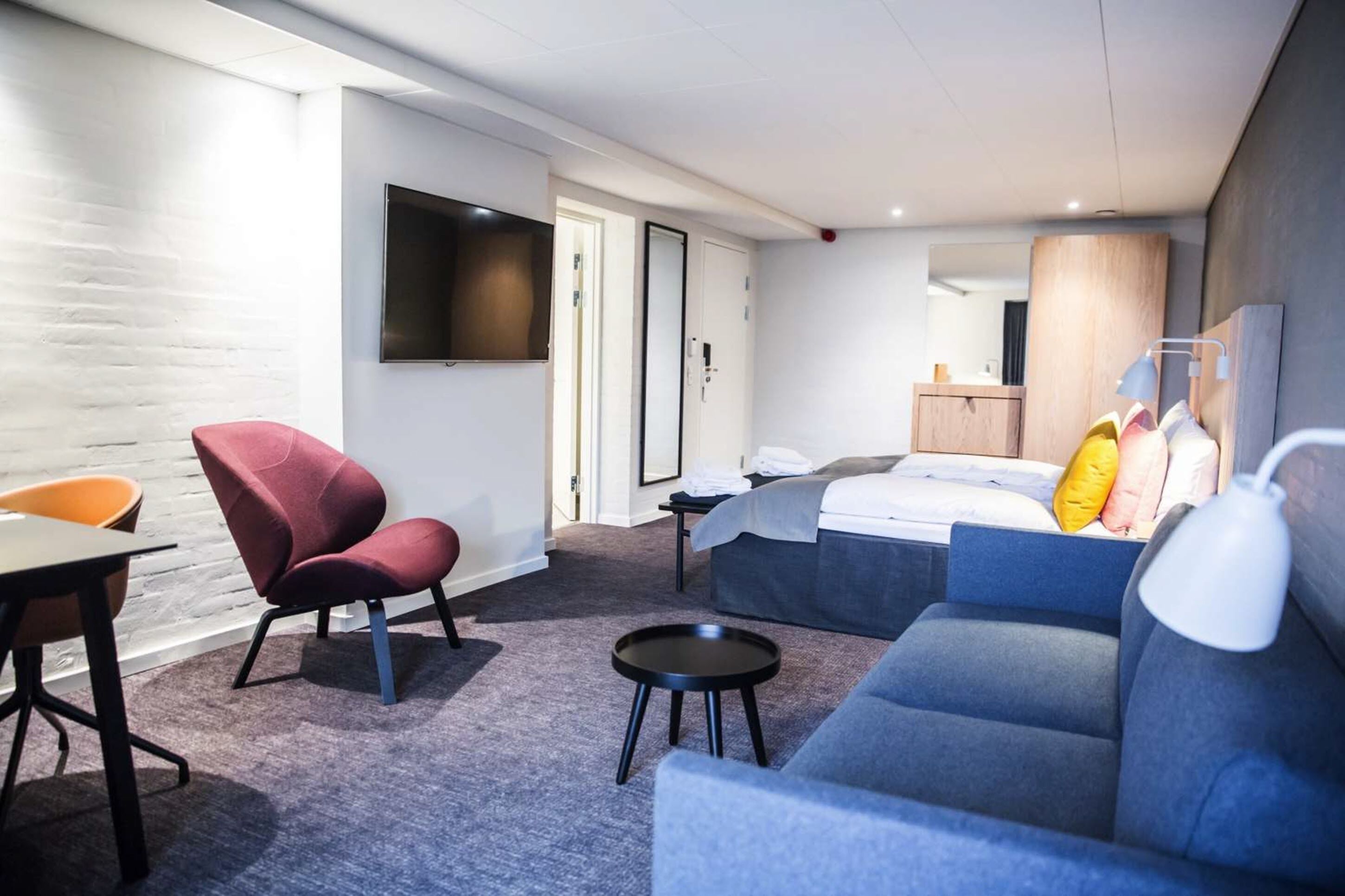Un hotel moderno con un sofá gris, un sillón rojo, una pequeña mesa redonda, una cama con cabecera gris y un televisor de pantalla plana montado en la pared.