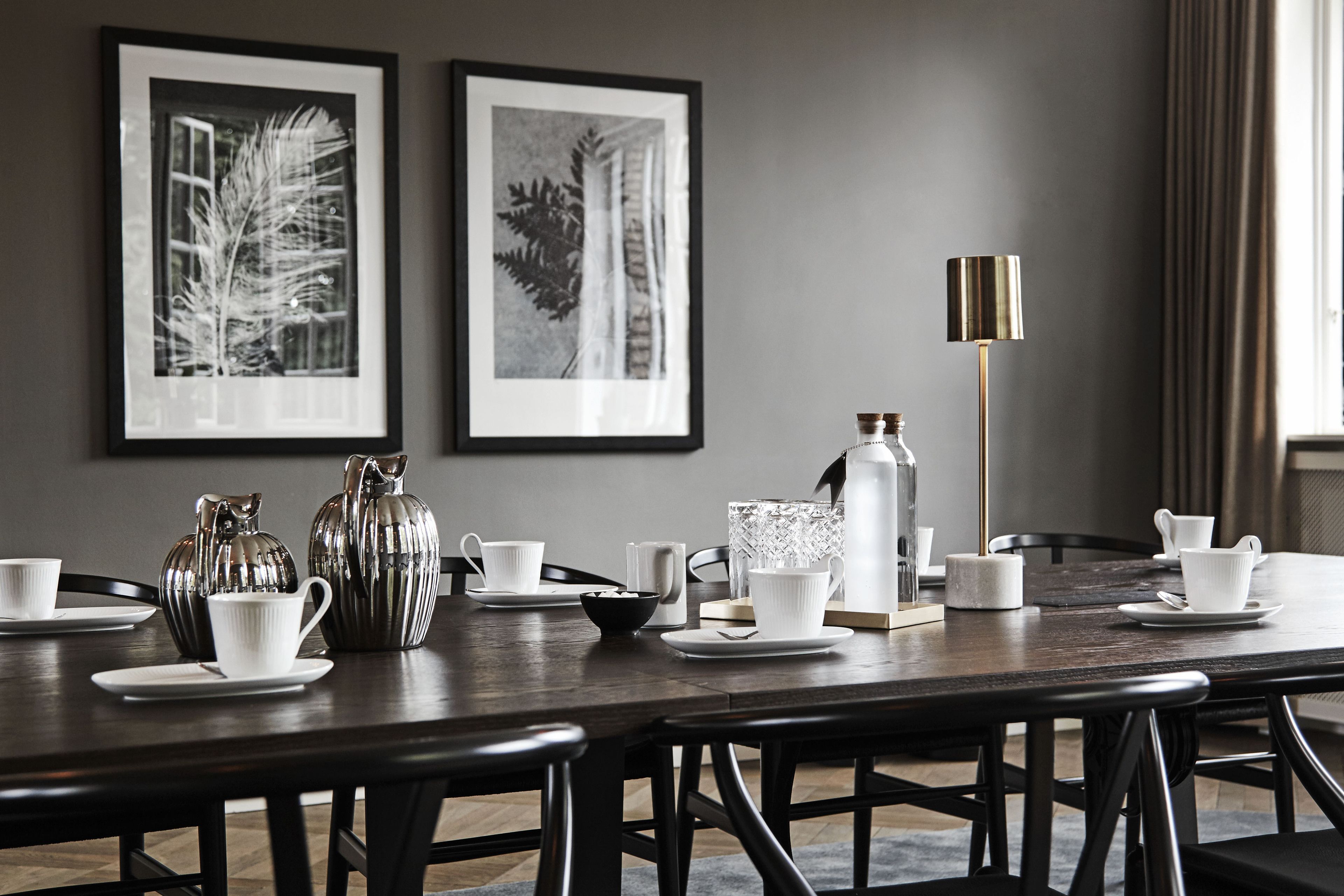 Una mesa de comedor dispuesta con tazas, platillos y una jarrón plateado, con arte blanco y negro en la pared y una lámpara de pie alta.