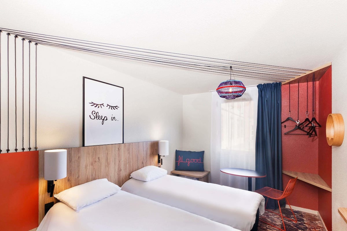 A hotel room with two beds, a wall-mounted picture, a hanging light fixture, a small table with a chair, and a red wall.