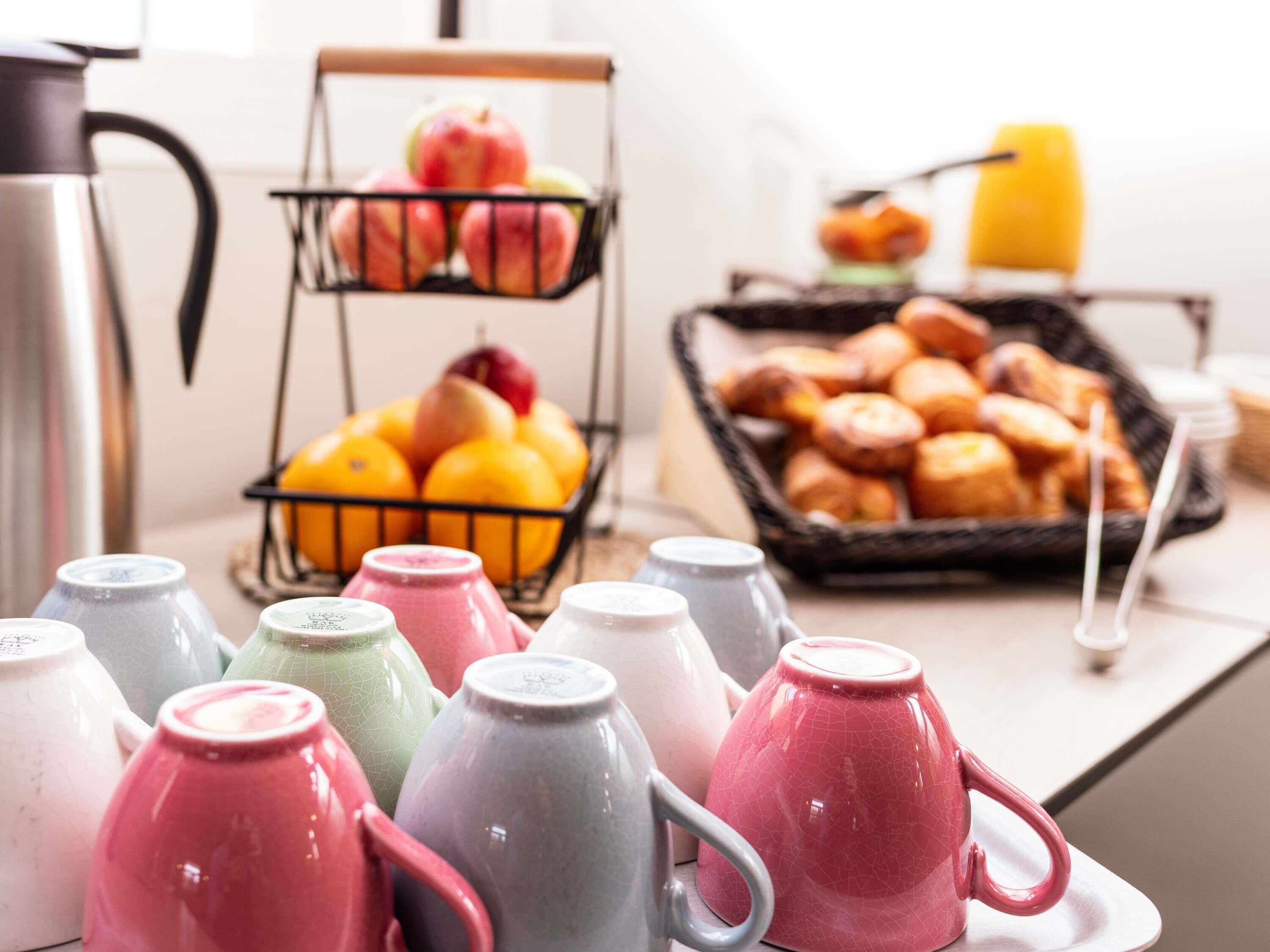 A breakfast setup with mugs, fruits, pastries, and a kettle.