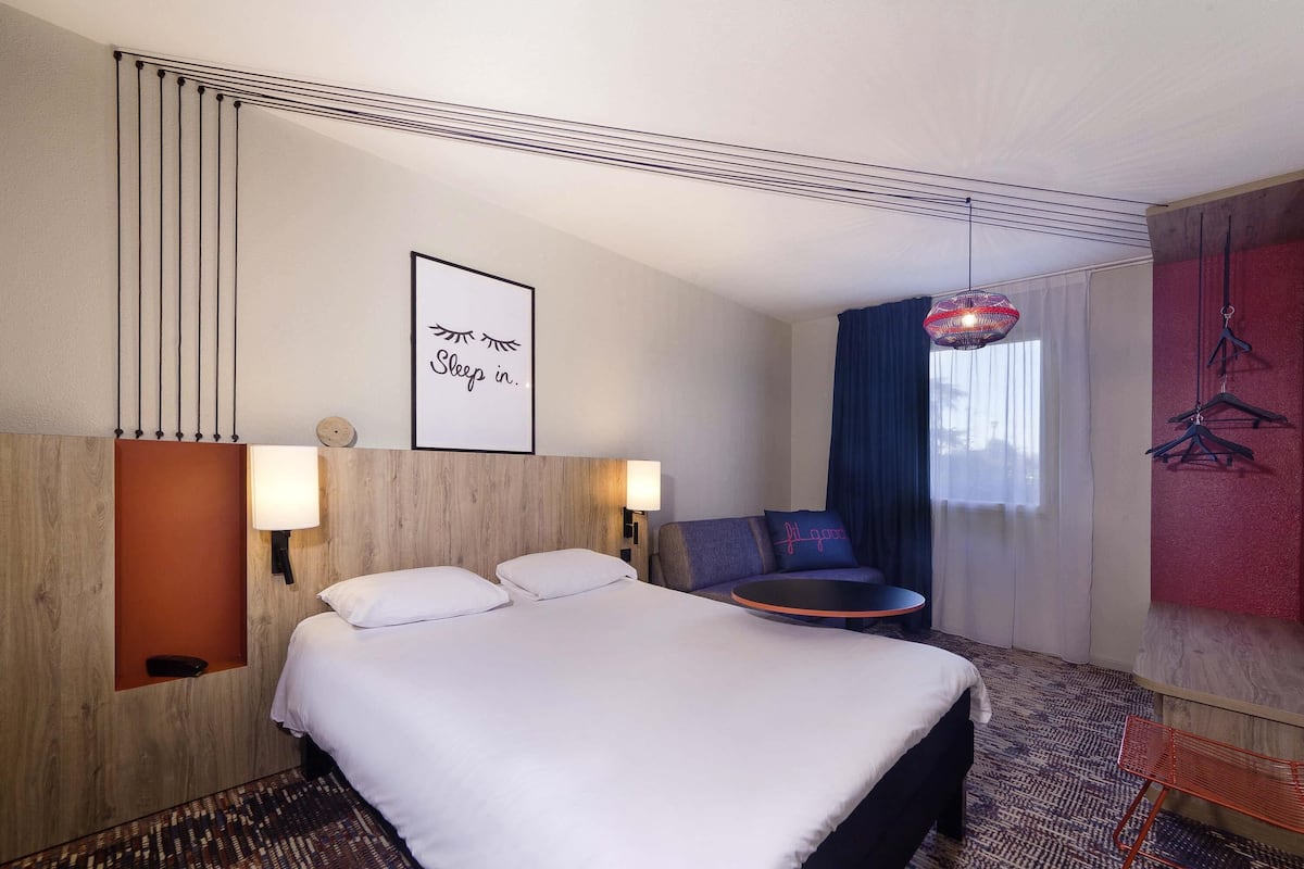 A hotel room with a bed, two bedside lamps, a wall-mounted artwork, a sofa, and a hanging light fixture.