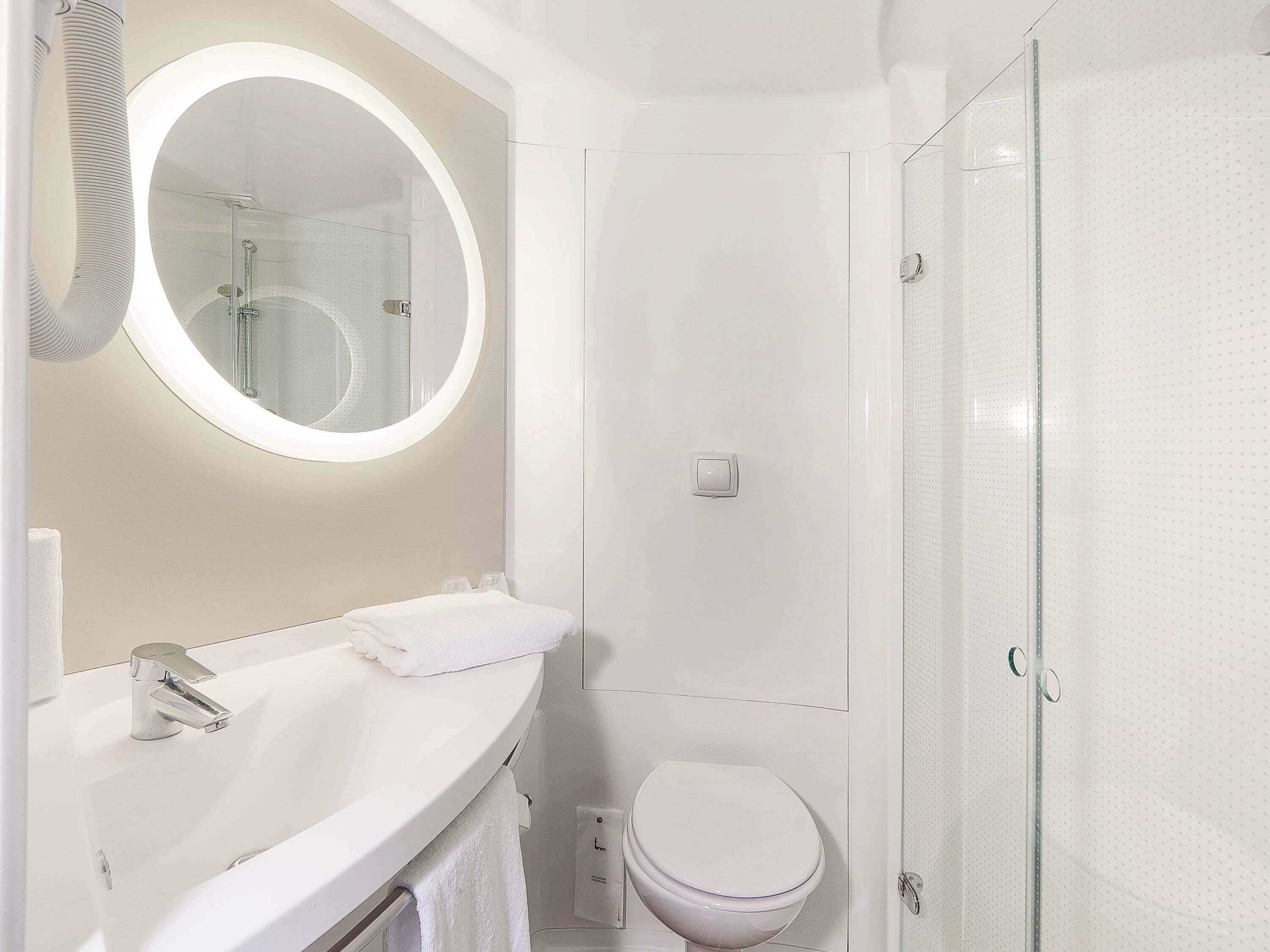 A modern bathroom with a round mirror, a white sink, a toilet, and a glass shower enclosure.