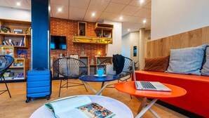 Un espace de détente moderne avec un mur en briques, un canapé rouge, une valise bleue et un ordinateur portable posé sur une petite table.