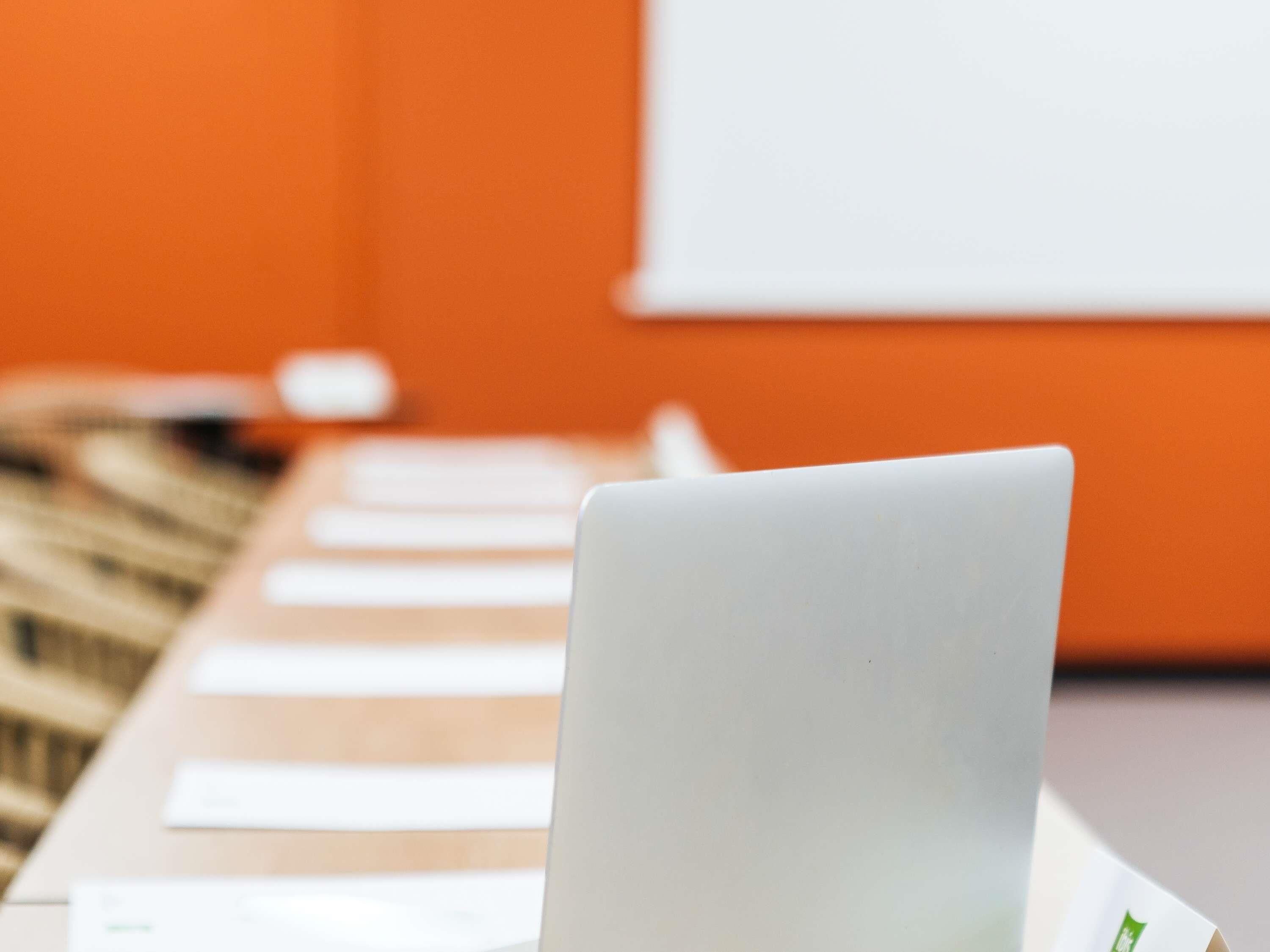 A laptop on a table with papers, a whiteboard, and a patterned rug in the background.