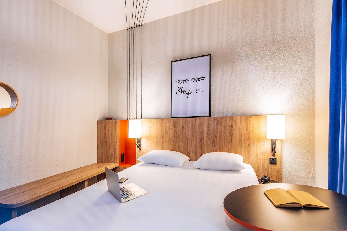 A hotel room with a bed, two bedside lamps, a laptop, a book, and a framed sign on the wall.