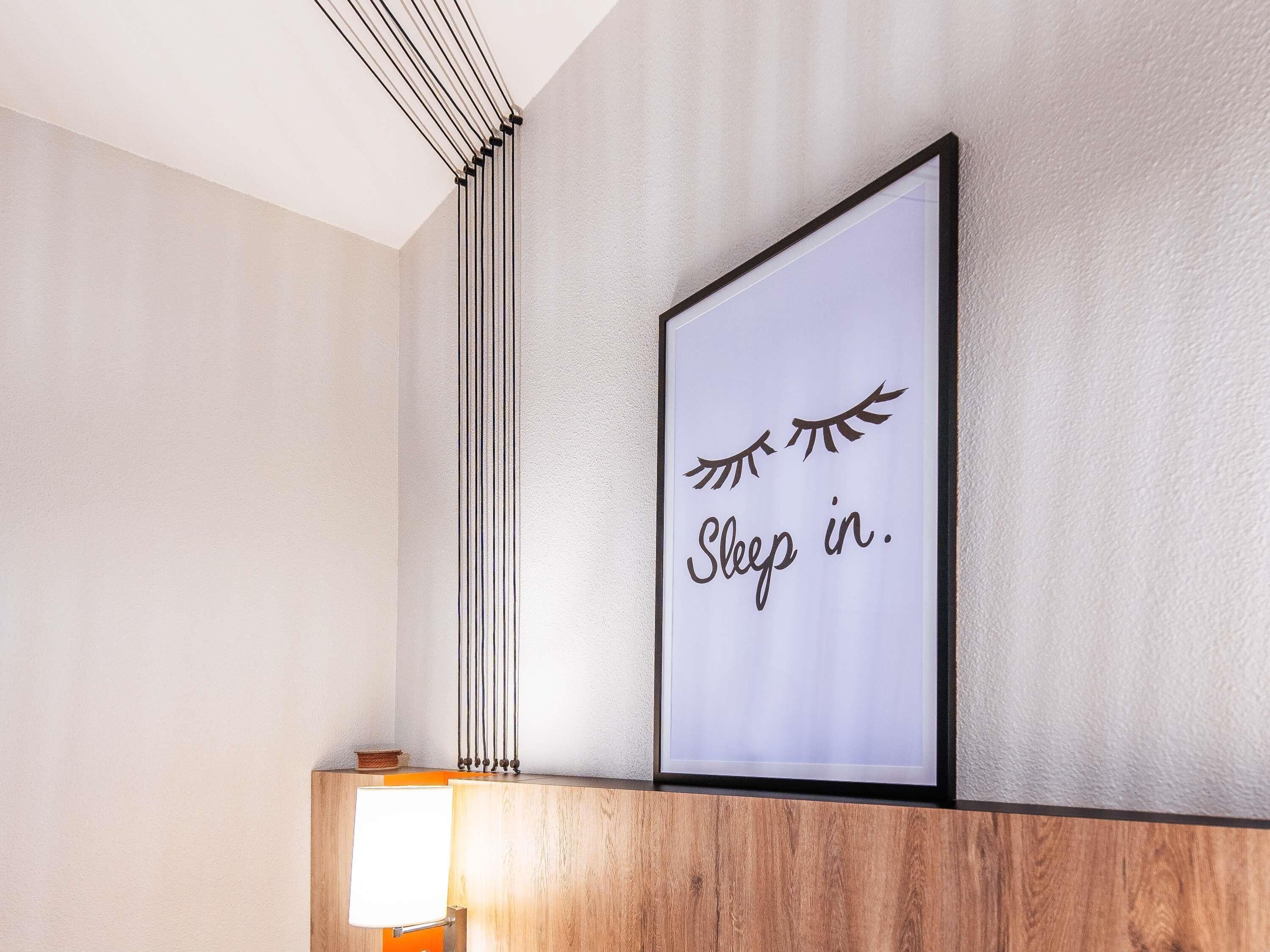 A framed sign with the text 'Sleep in.' on a wall, a wooden shelf below, and a lamp to the left.