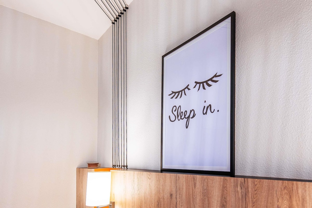 A framed sign with the text 'Sleep in.' on a wall, a wooden shelf below, and a lamp to the left.