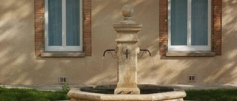 A stone fountain with a spherical top, set in a circular stone basin, in front of a building with two windows and brick arches.