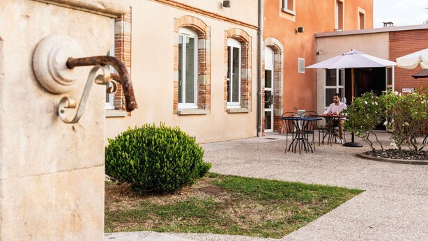 Une cour avec une table et des chaises, un petit arbuste et un bâtiment à la façade en briques et aux fenêtres en arc de cercle.