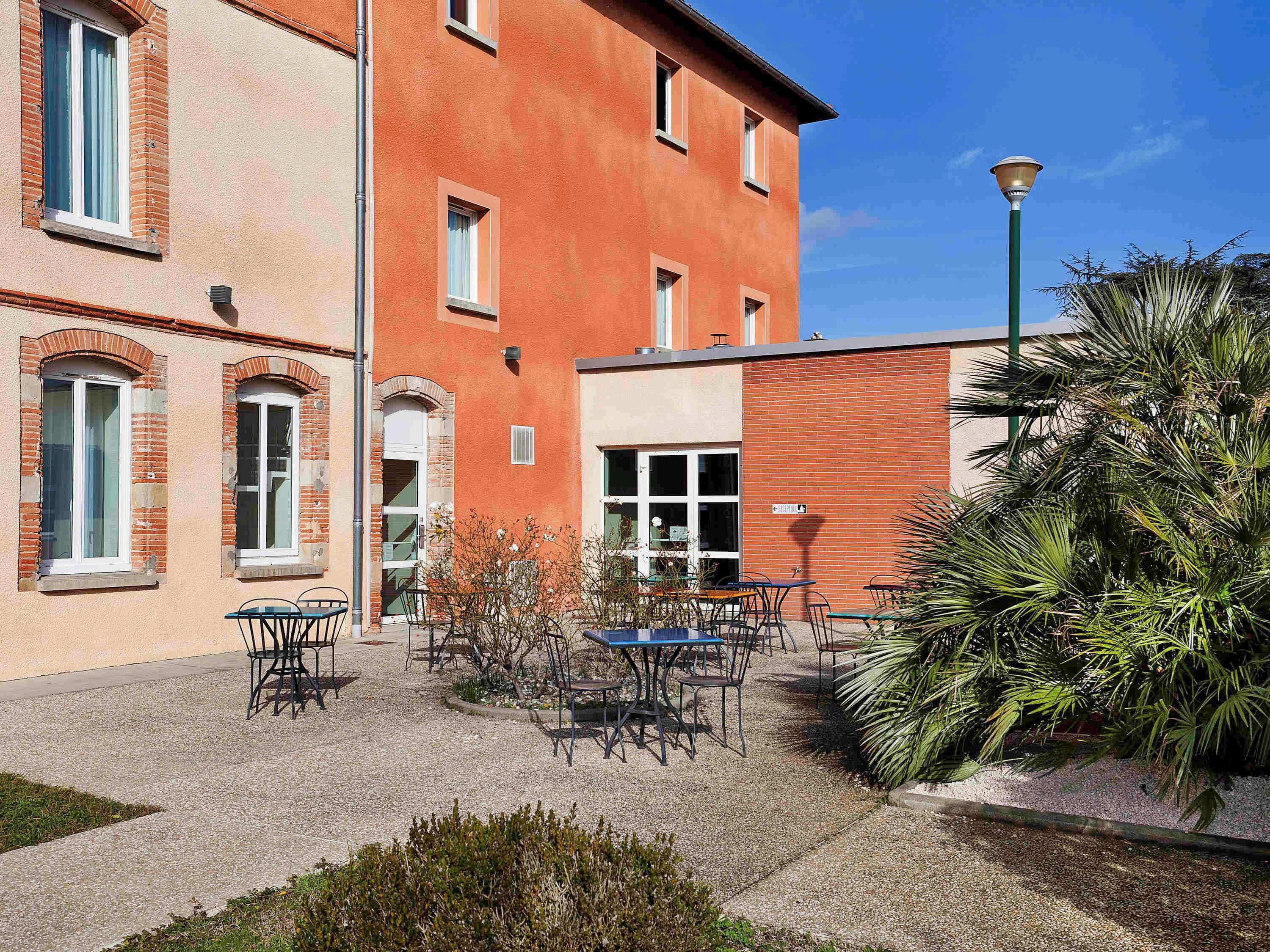 Un bâtiment à la façade rouge, doté de nombreuses fenêtres et d’un espace extérieur aménagé avec des tables et des chaises.