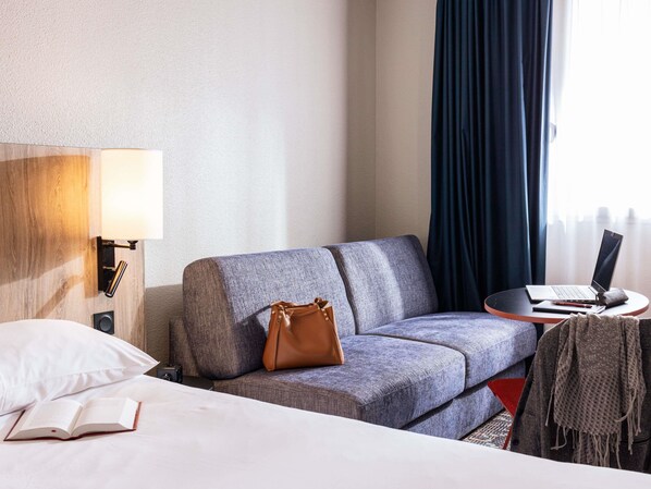Une chambre d’hôtel avec un lit, un canapé, un bureau avec un ordinateur portable, un sac à main et une lampe.