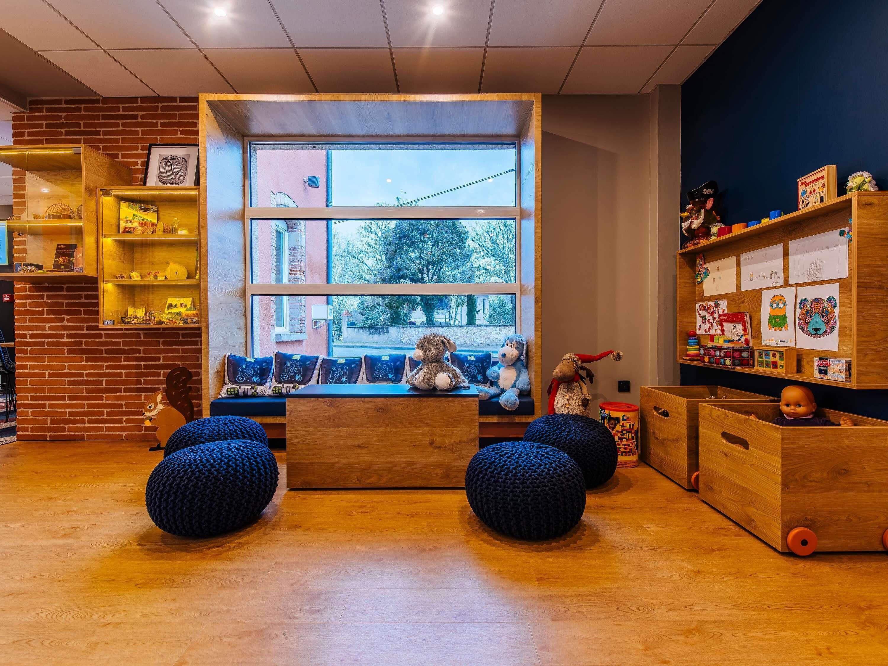 A cozy room with a large window, a wooden bench with cushions, and a bookshelf filled with children's books and toys.