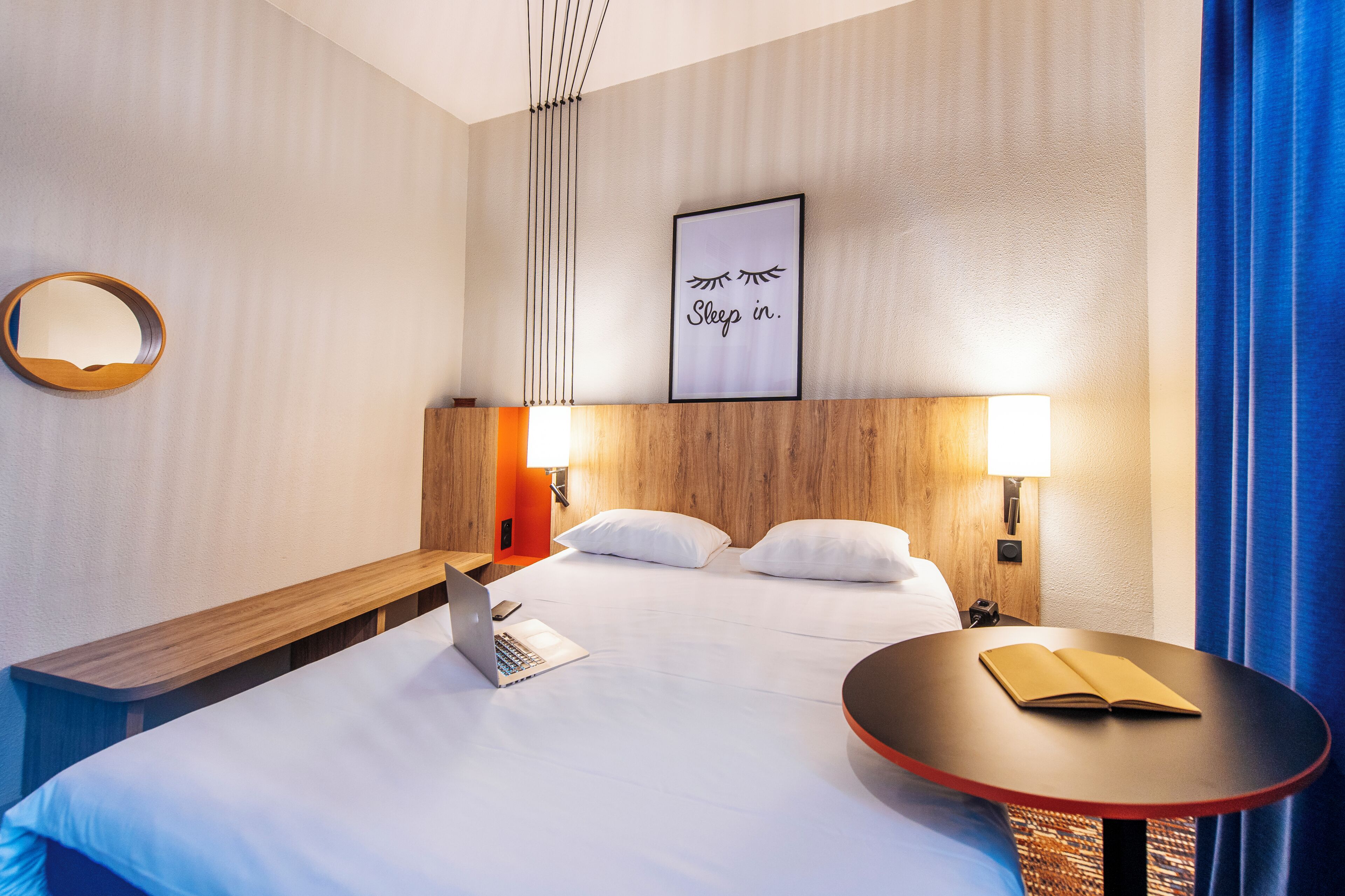 A hotel room with a bed, a desk with a laptop, a round table with a book, a wall-mounted mirror, and a framed sign on the wall.
