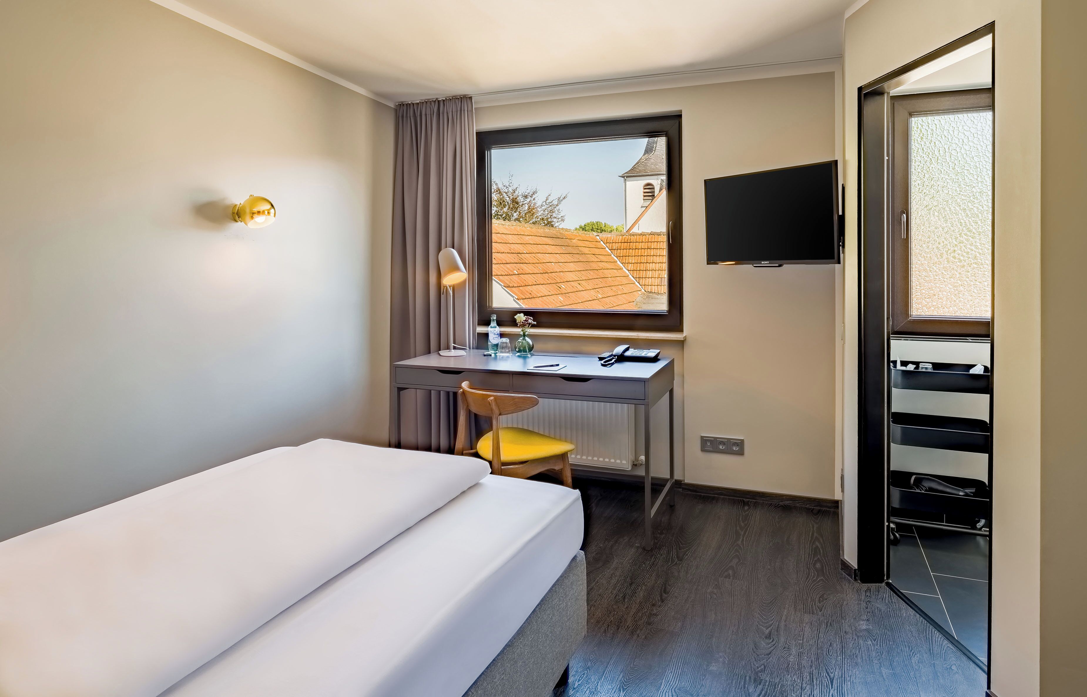 Quarto de hotel com cama, escrivaninha, cadeira e televisão.
